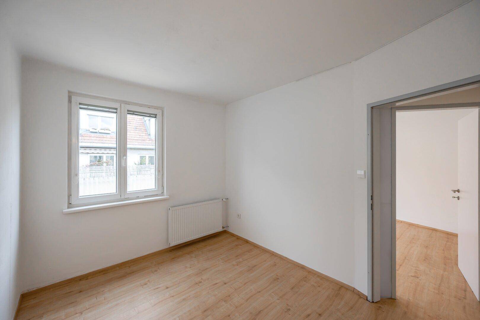 Leeres Zimmer mit Laminatboden, großem Fenster und Blick auf die umliegenden Dächer.
