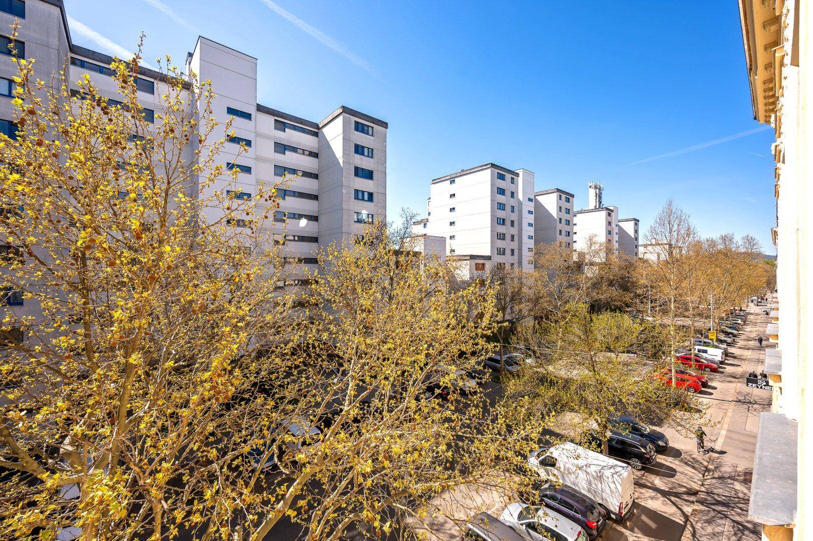 Weitblick von einem Balkon über die Stadt mit Bäumen und Wohngebäuden unter blauem Himmel.