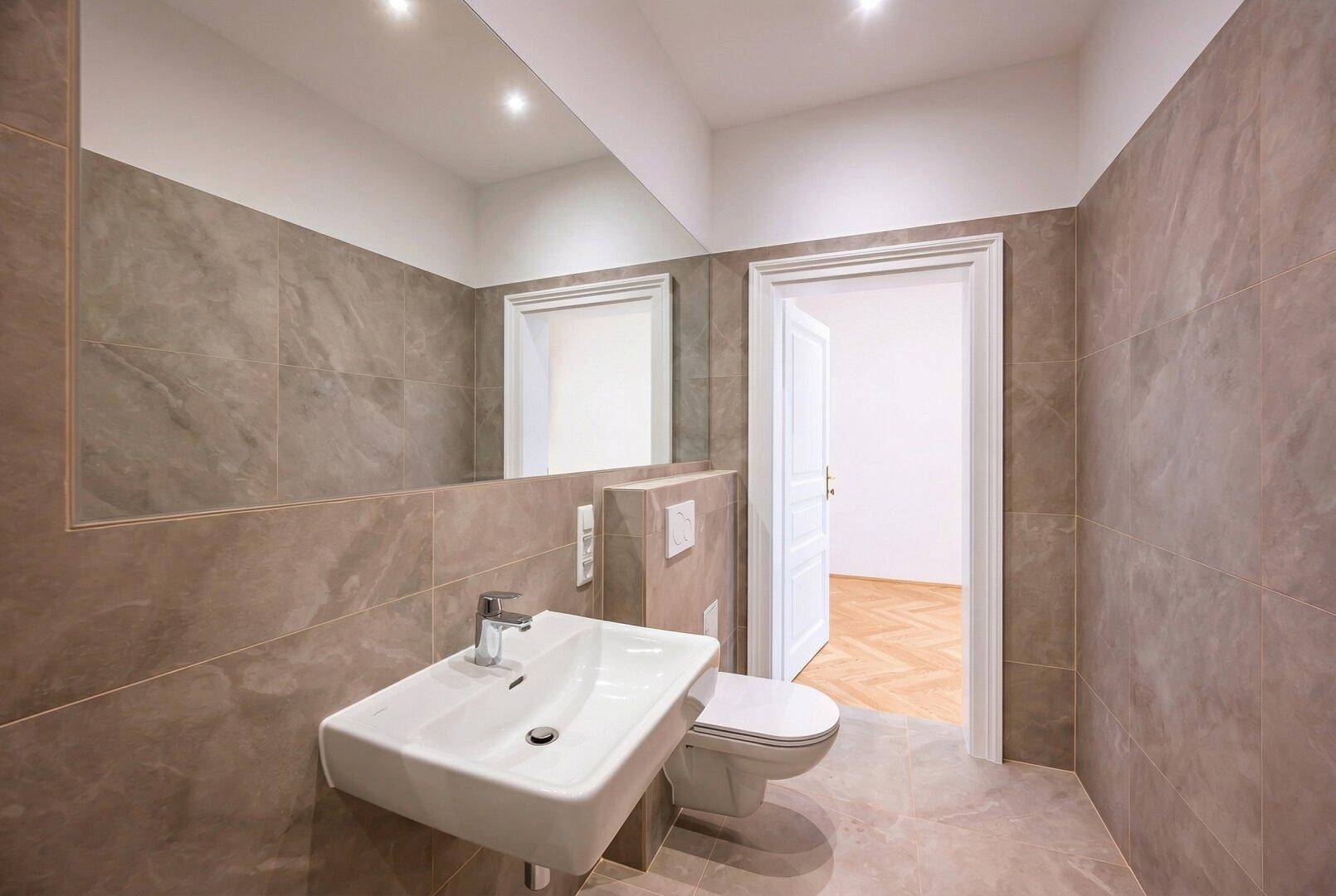 Helles Badezimmer mit großformatigen Fliesen, wandhängendem WC und Blick in den Flur.