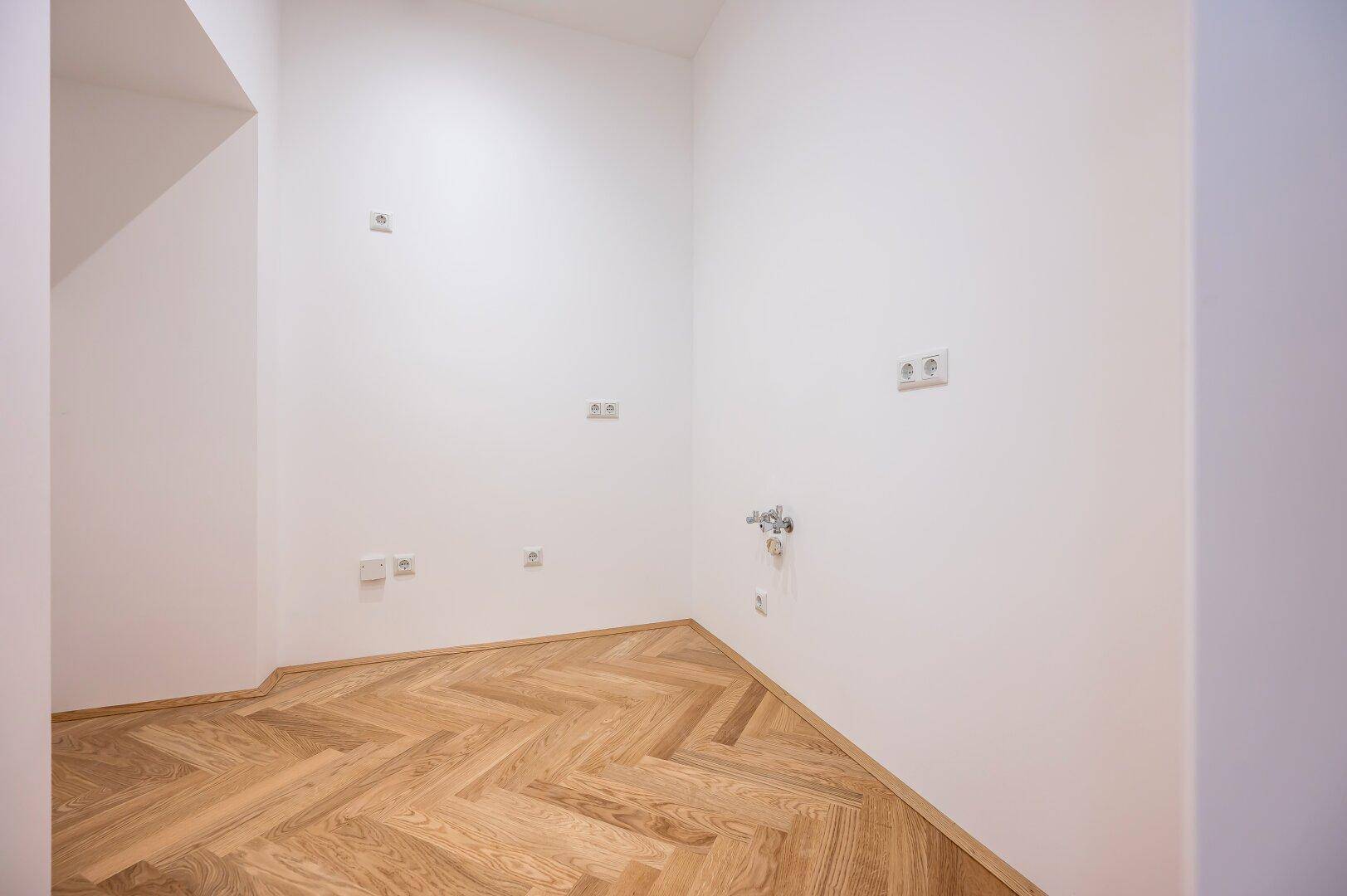 Leerer Raum mit Anschlüssen für eine Küche und Fischgrätparkett, bereit für den Einbau.