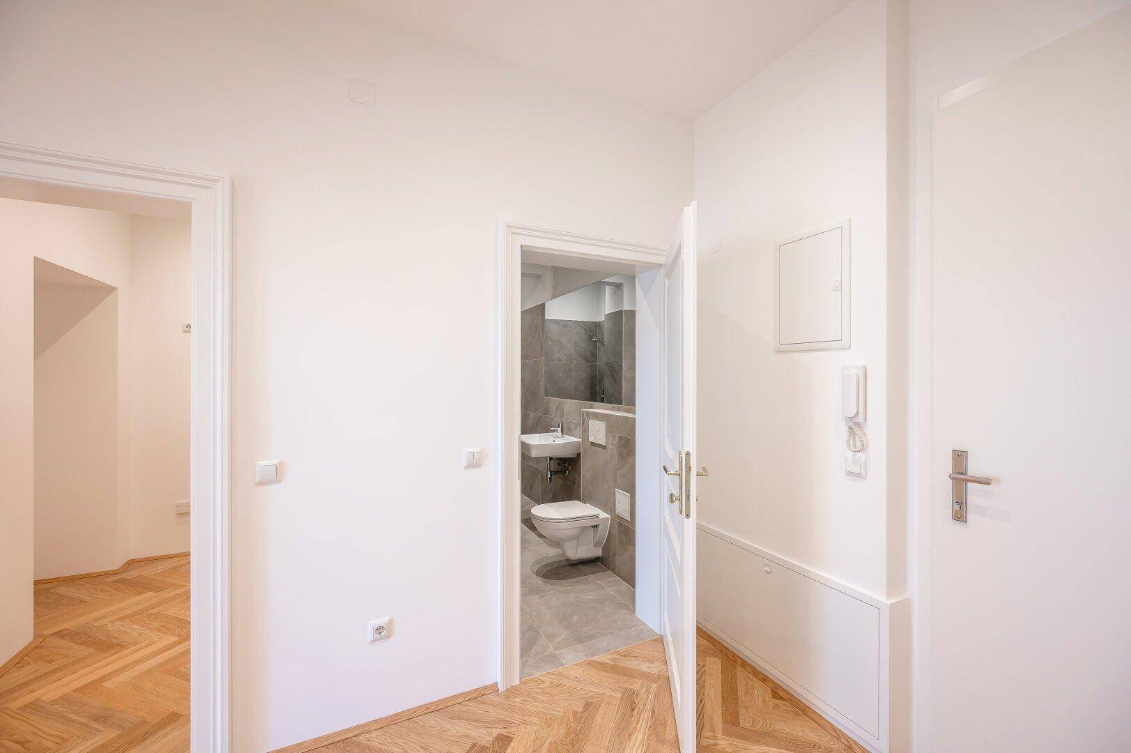 Eingangsbereich mit Fischgrätparkett und offener Tür zu einem modernen Badezimmer.