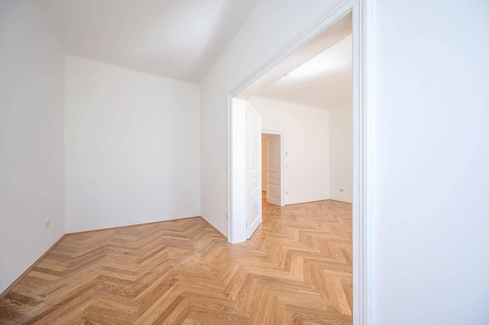 Geräumiger, heller Raum mit Fischgrätparkett und hohen Decken, ideal für ein Wohnzimmer.