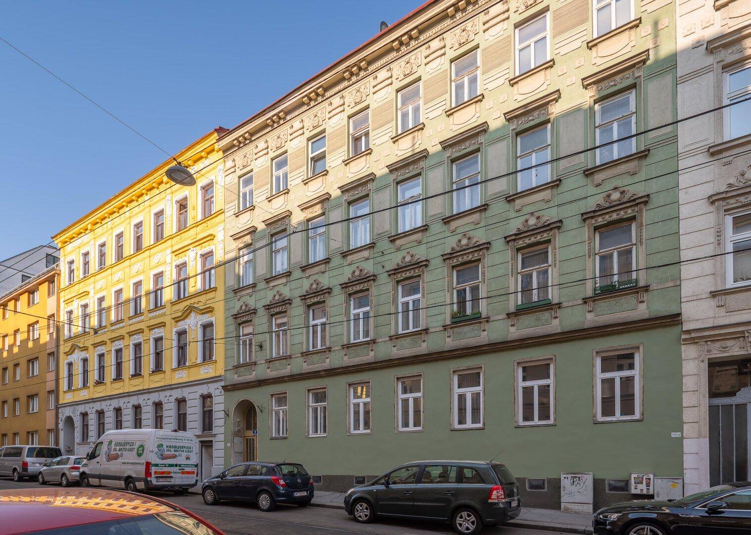 Historisches Mehrfamilienhaus mit grüner Fassade und vielen Fenstern an einer Stadtstraße.