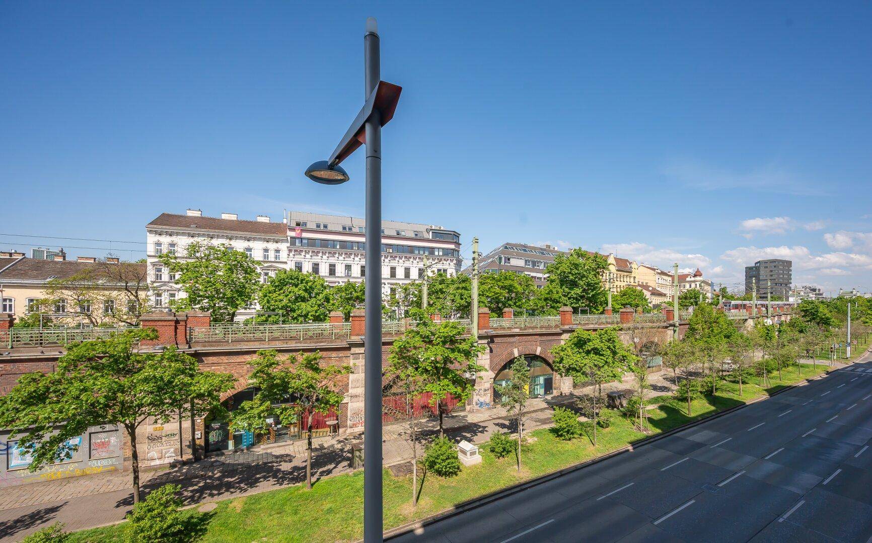 Weitläufige Straßenansicht mit Bäumen, Gebäuden und einer historischen Bogenbrücke unter blauem Himmel.