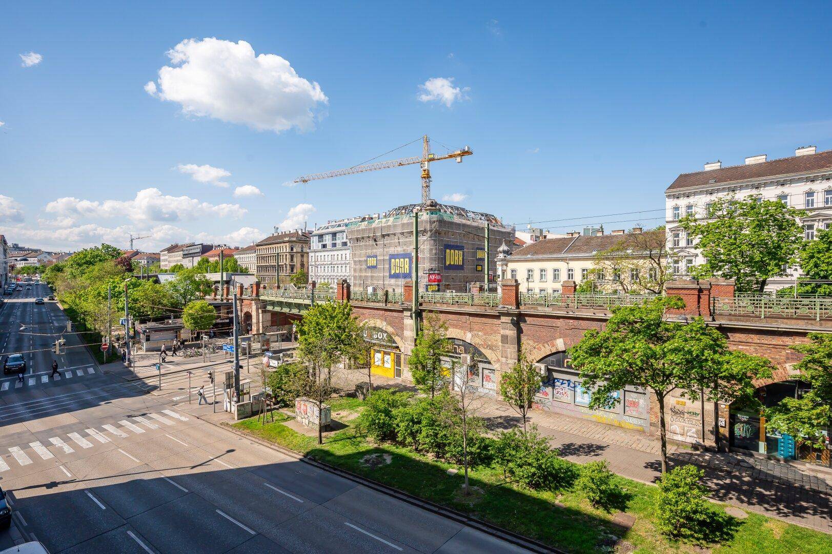 Panoramablick auf eine belebte Stadtstraße mit Gebäuden, Bäumen und einer Baustelle im Hintergrund.