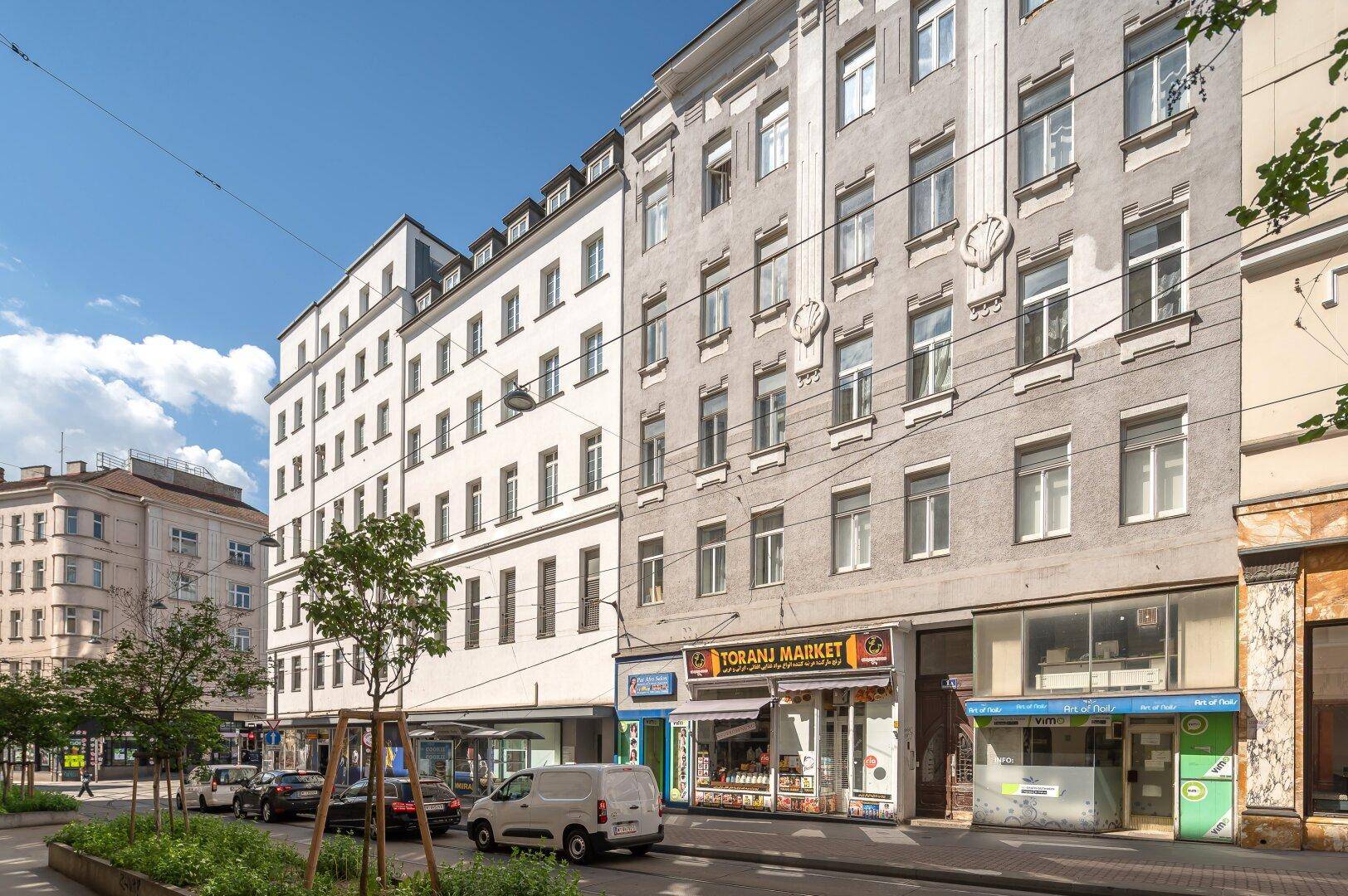 Fassade eines Altbaus mit Geschäften im Erdgeschoss und parkenden Autos an der Straße.