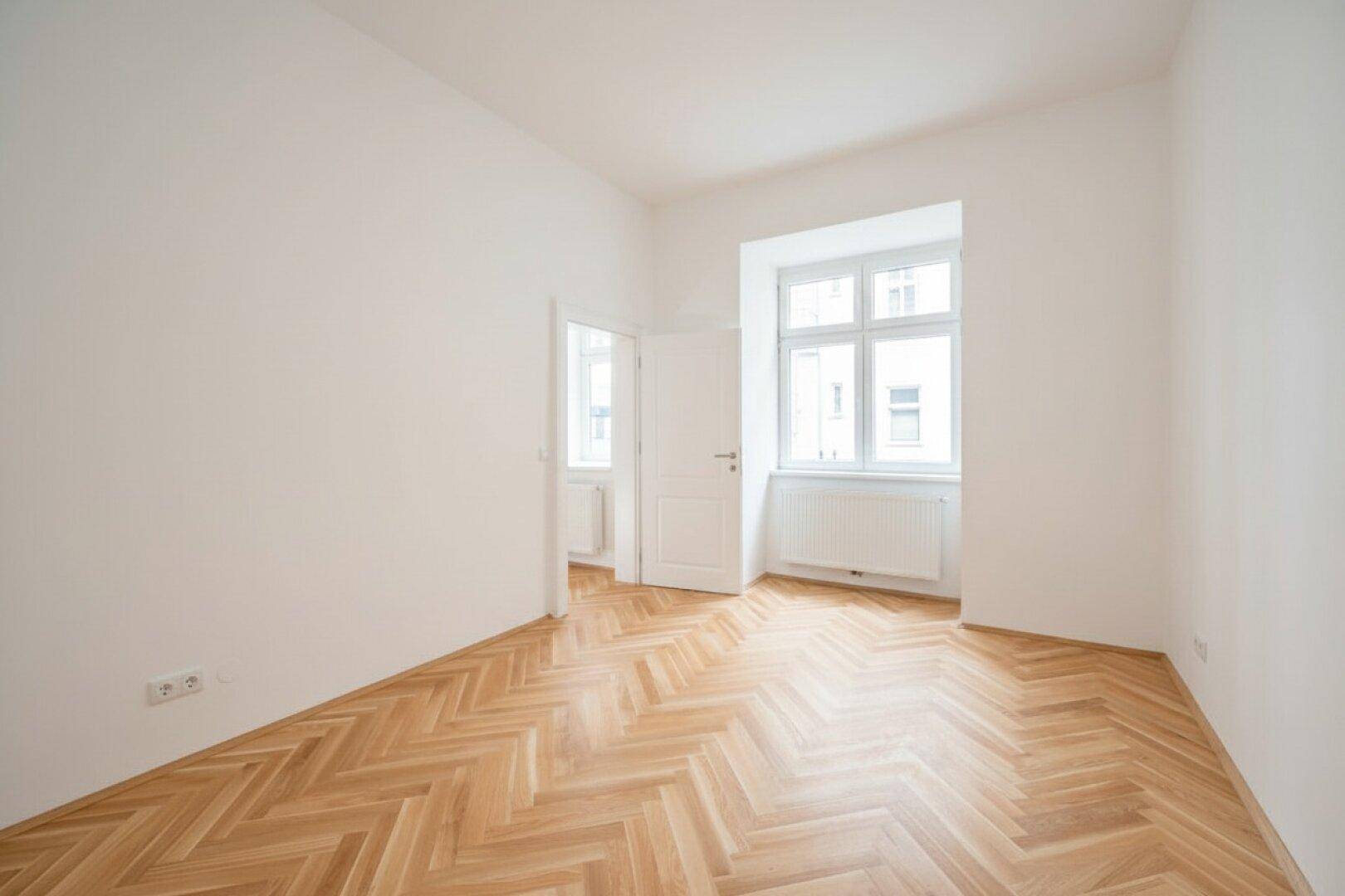 Lichtdurchflutetes Zimmer mit klassischem Fischgrätparkett und einem Fenster mit Heizkörper darunter.