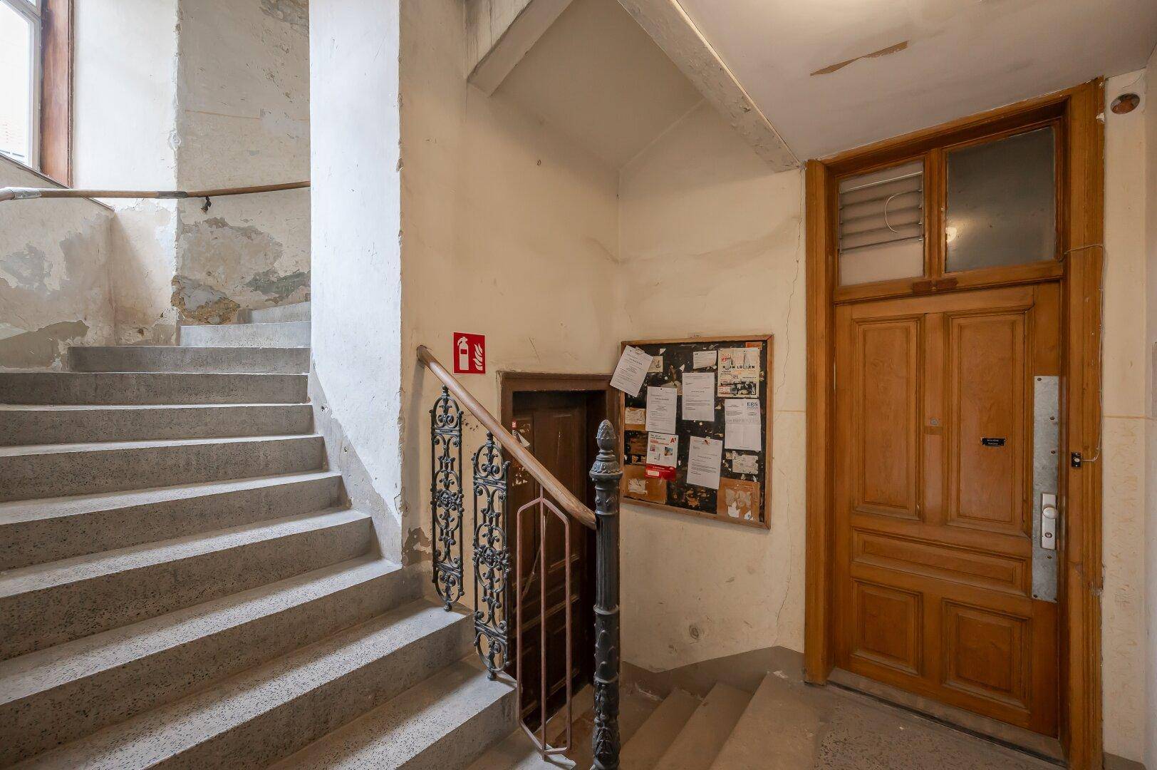 Historisches Treppenhaus mit Betonstufen, schmiedeeisernem Geländer und Holztür in einem Altbau.
