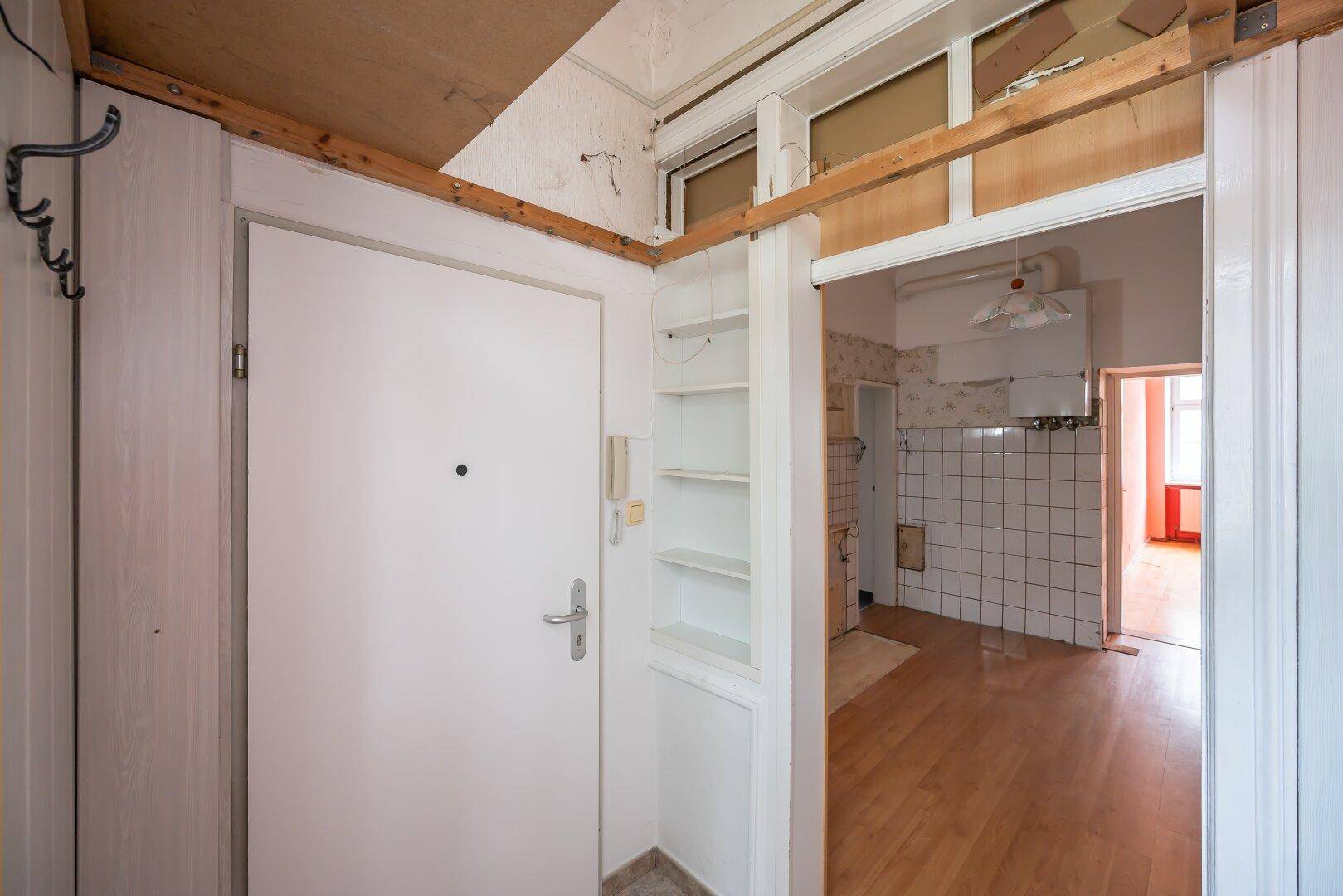 Eingangsbereich mit weißer Tür, eingebauten Regalen und Blick in die renovierungsbedürftige Küche.