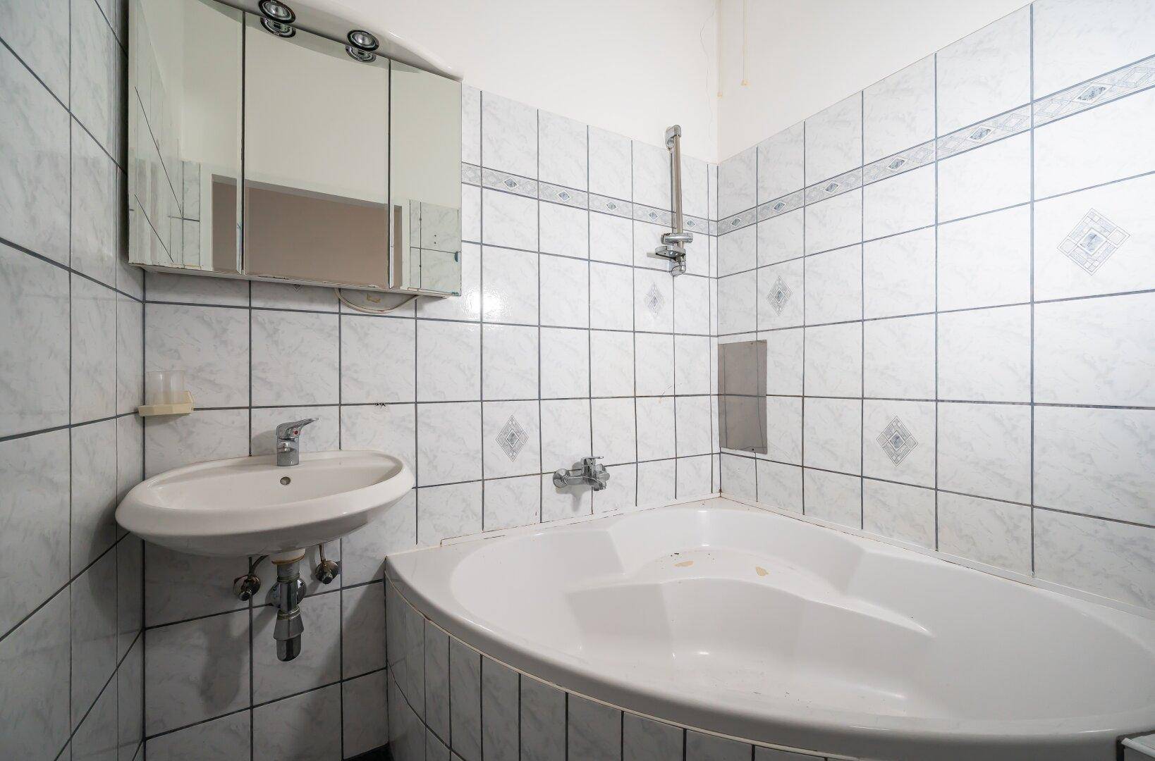 Badezimmer mit weißen Fliesen, Eckbadewanne, Einzelwaschbecken und Spiegelschrank.