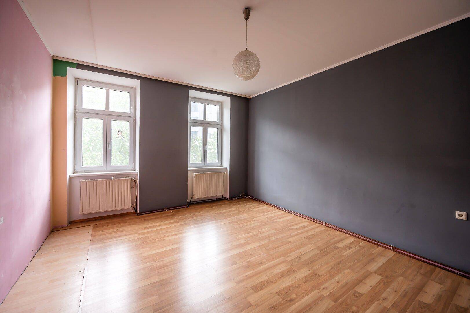 Geräumiges Zimmer mit dunklen und farbigen Wänden, Holzboden und zwei Fenstern, bietet viel Gestaltungspotenzial.