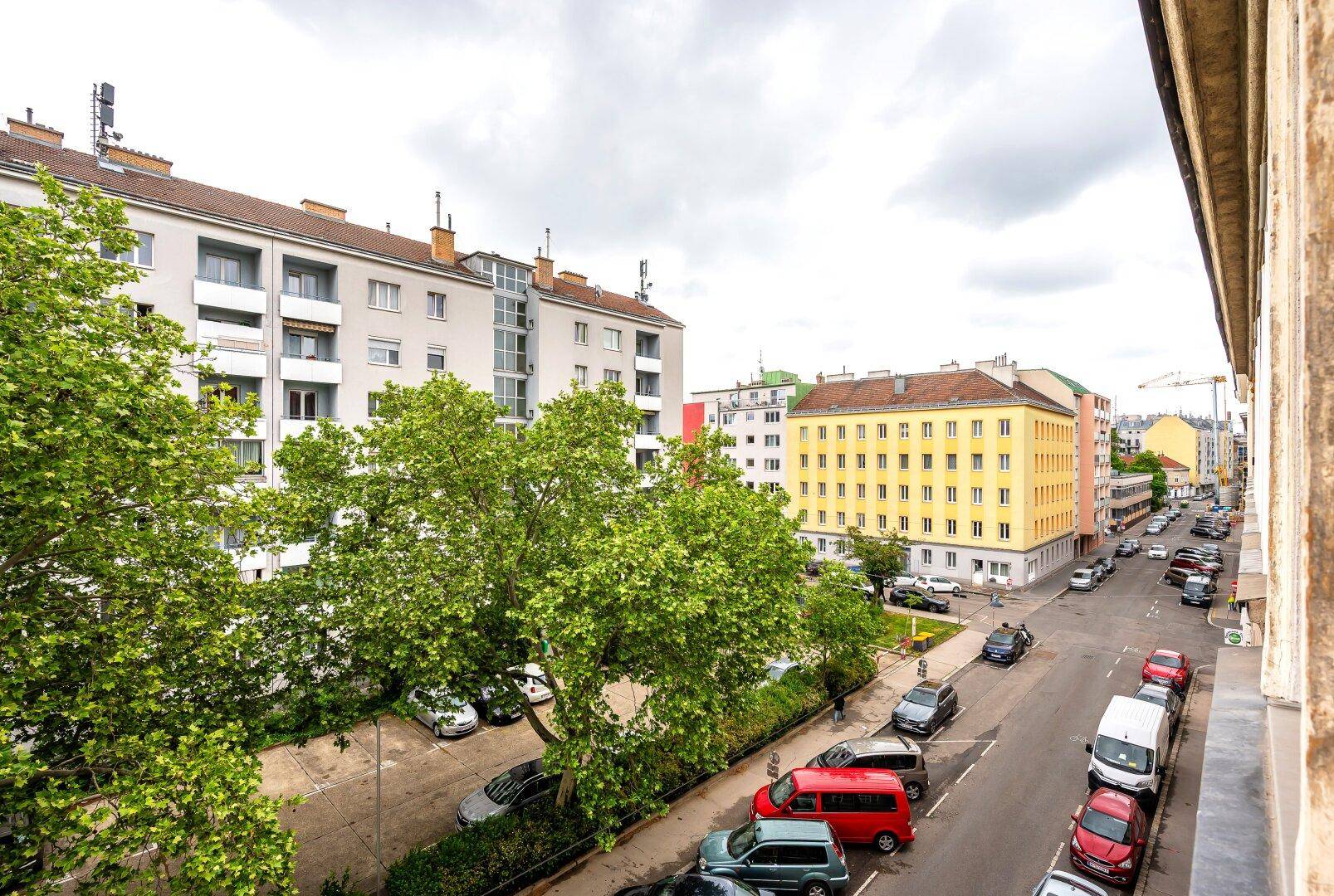 Blick auf eine belebte Stadtstraße mit parkenden Autos, Bäumen und Wohngebäuden.