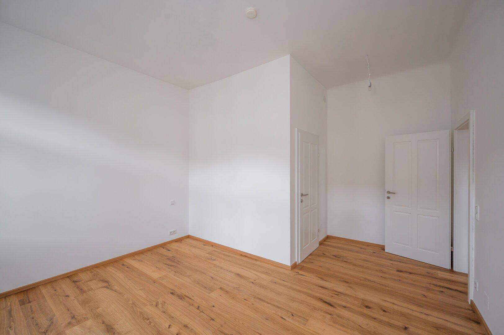 Leeres Zimmer mit Holzboden und zwei Türen, bietet Zugang zu weiteren Räumen.