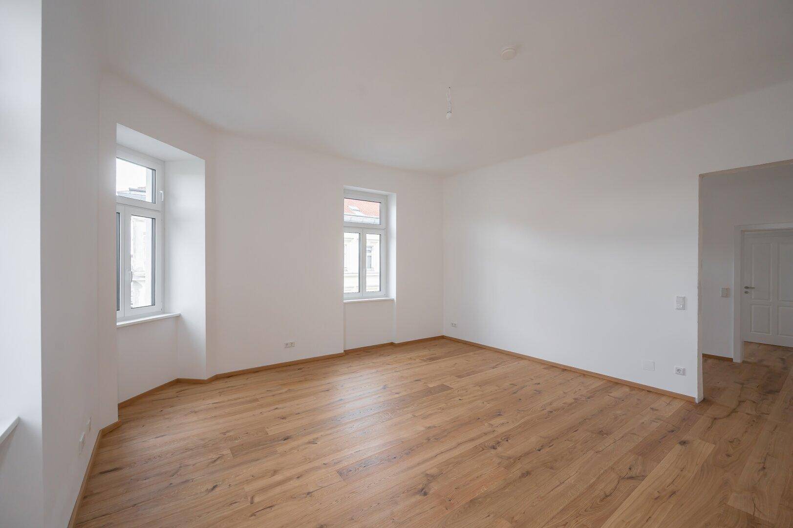 Heller, leerer Raum mit Holzboden und zwei Fenstern, ideal für Wohn- oder Schlafzimmer.
