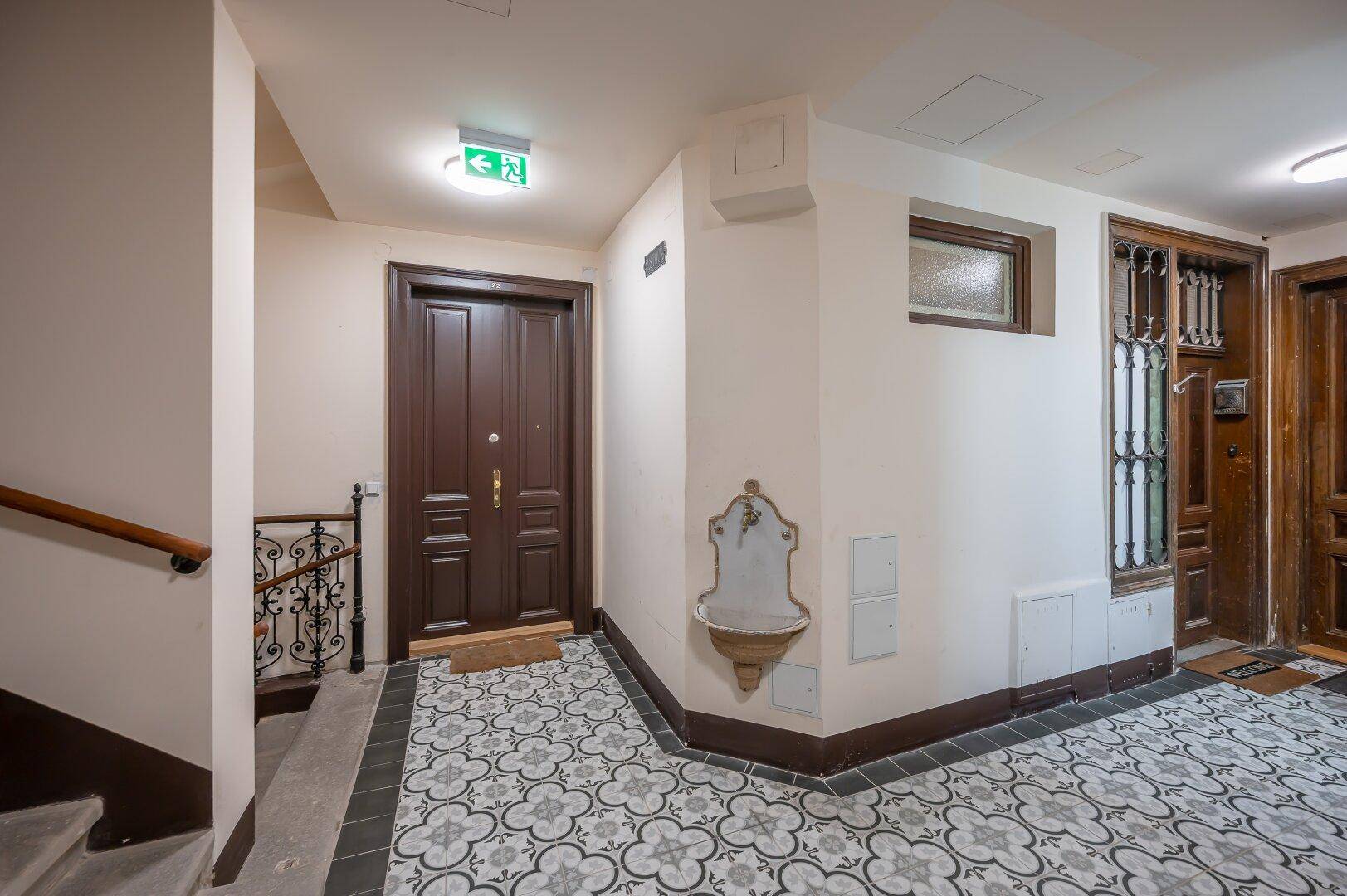 Historischer Eingangsbereich mit gemusterten Fliesen, Treppe und dekorativem Wandbrunnen.