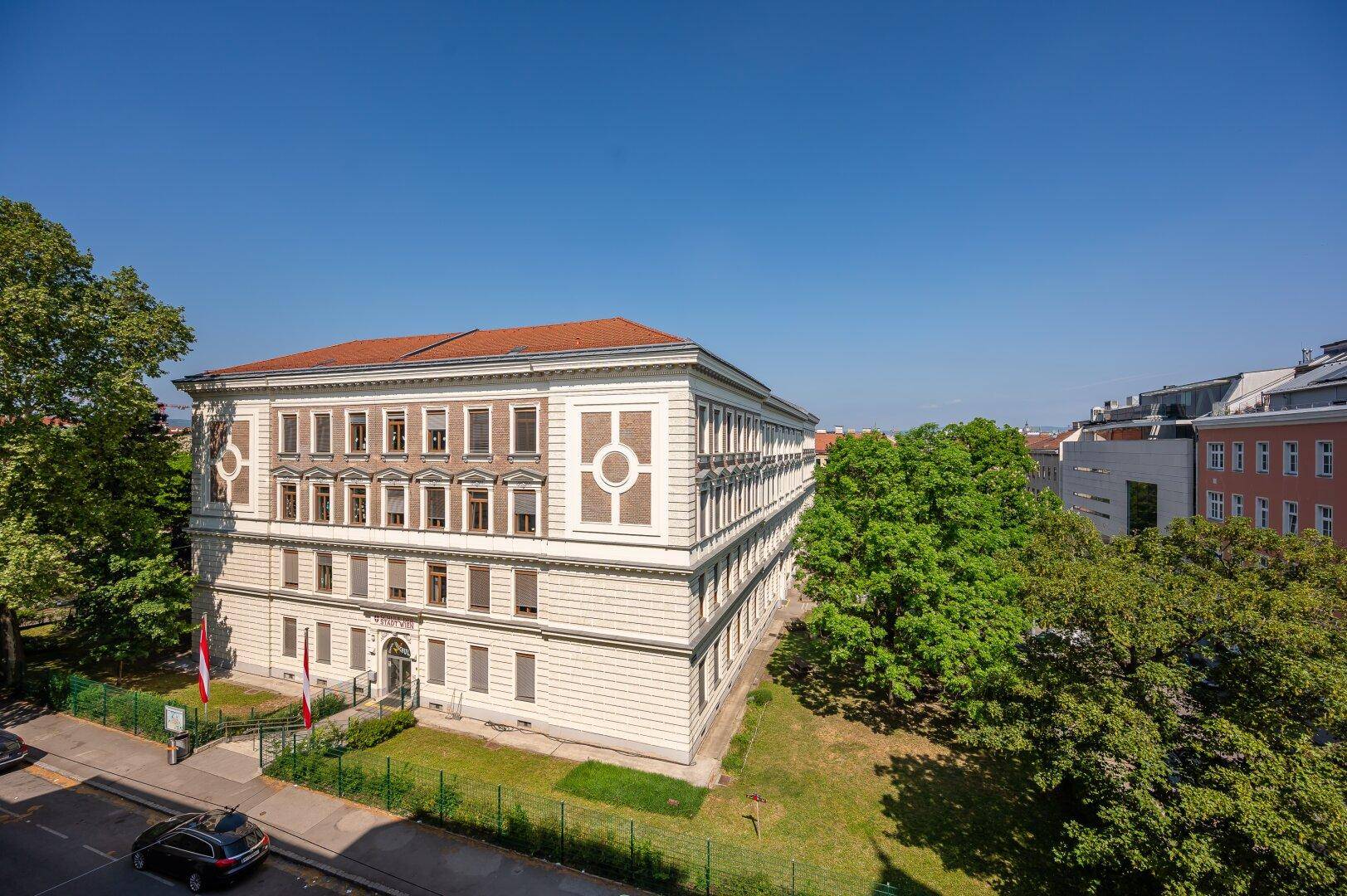 Historisches Gebäude mit klassischer Architektur, umgeben von Bäumen und Grünflächen.