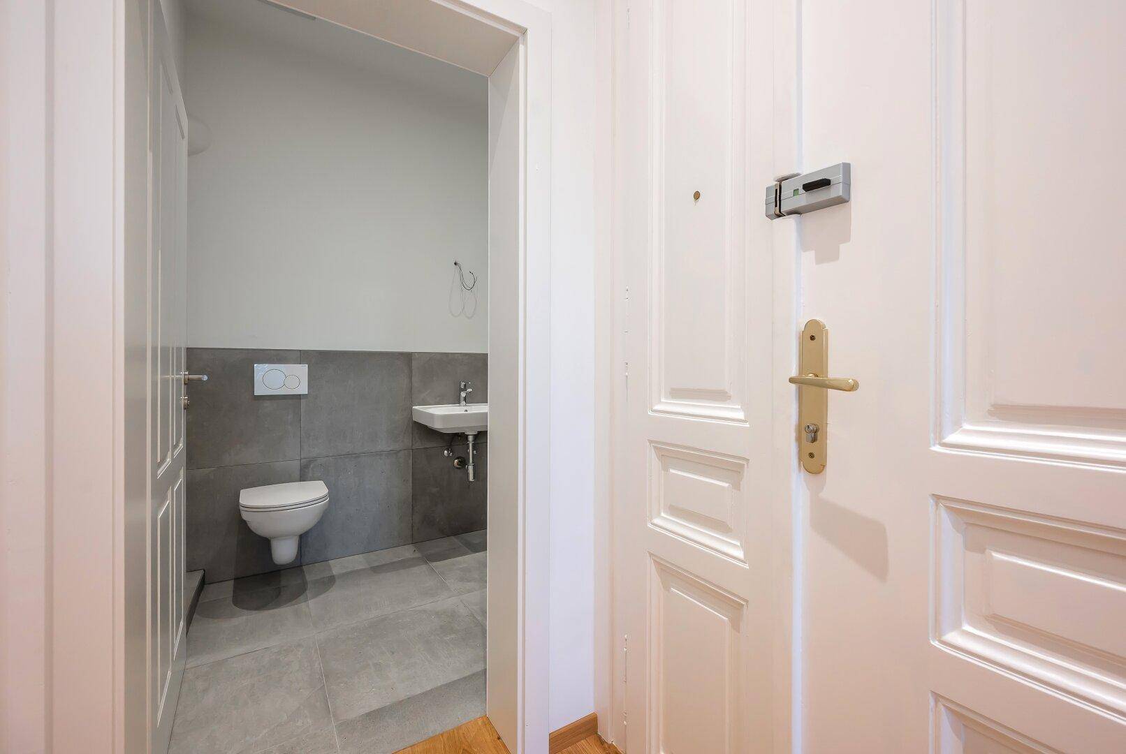 Modernes Gäste-WC mit Wand-WC, Waschbecken und grauen Fliesen, direkt vom Flur zugänglich.