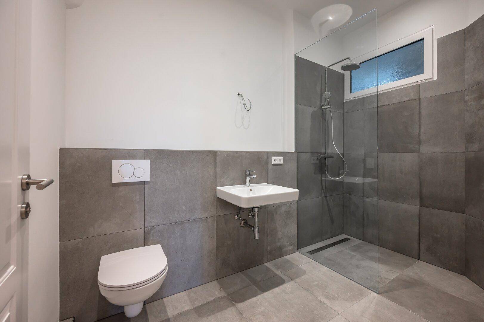 Helles Badezimmer mit ebenerdiger Dusche, Wand-WC, Waschbecken und Fenster für Tageslicht.
