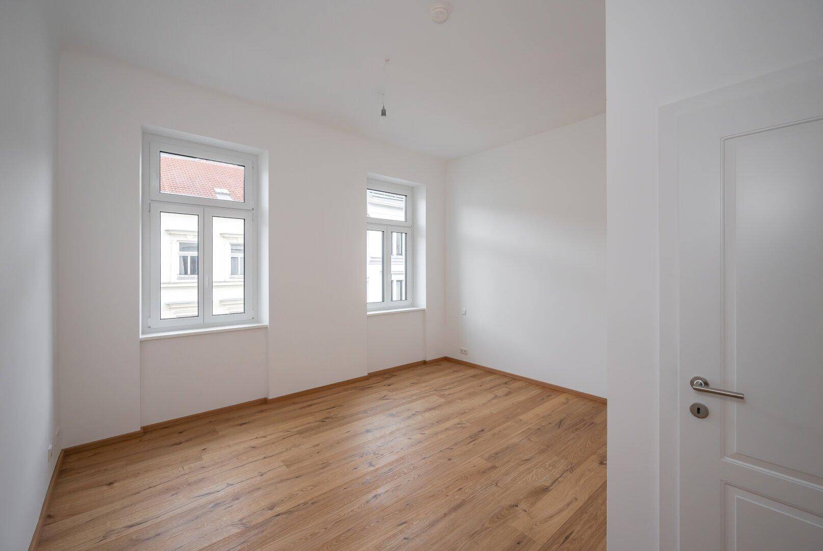Geräumiges, helles Zimmer mit Holzboden und zwei Fenstern, ideal als Schlaf- oder Wohnbereich.