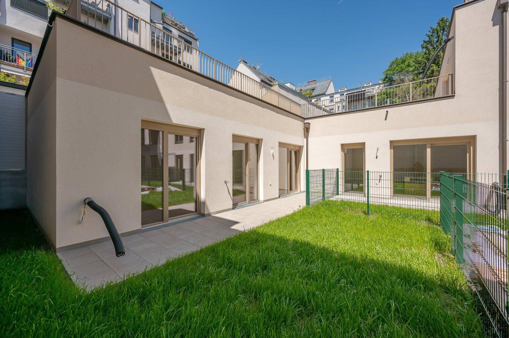 Privater Gartenbereich mit Rasenfläche und gepflasterter Terrasse, umgeben von modernen Gebäuden.