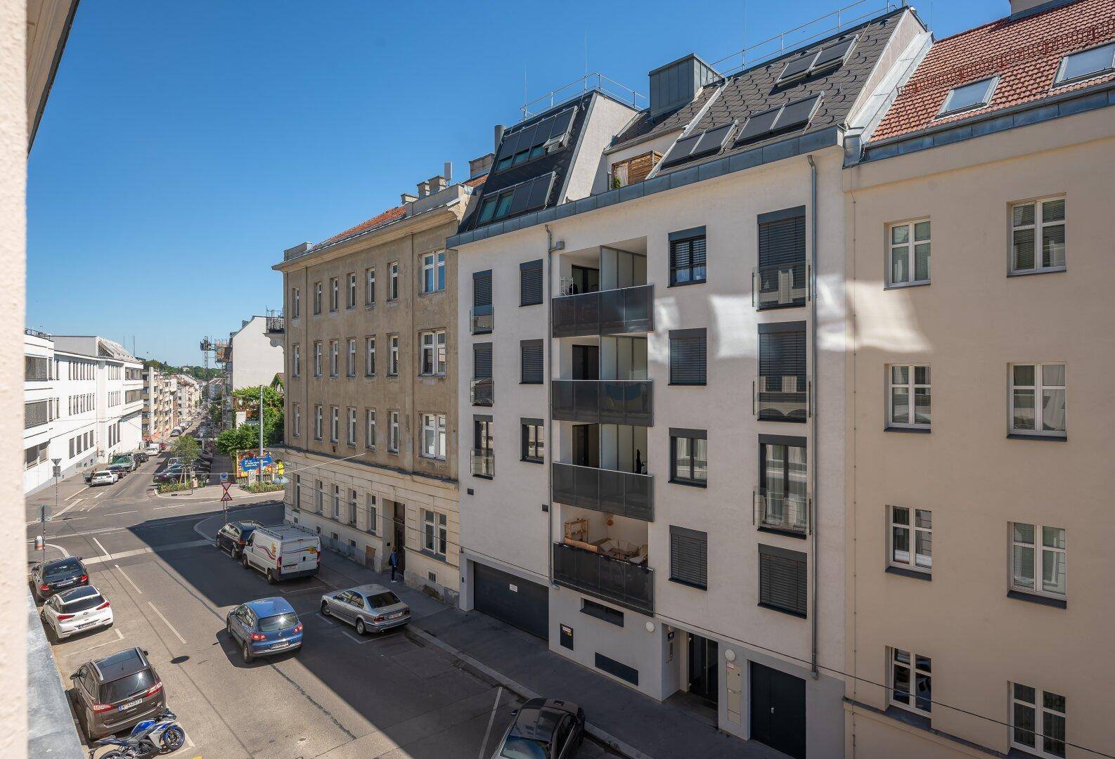 Blick von einem Balkon auf eine belebte Stadtstraße mit parkenden Autos und umliegenden Gebäuden.