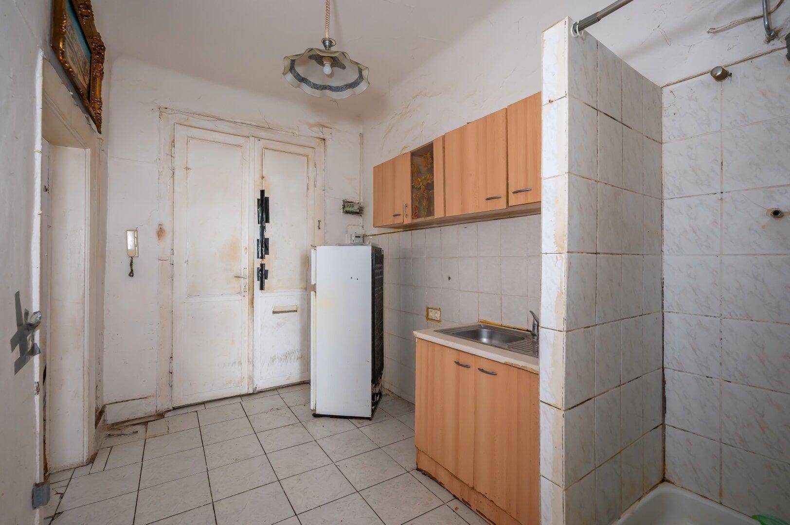 Kleine, einfache Küche mit Spüle, Kühlschrank und angrenzendem Duschbereich, alles in sehr altem Zustand.
