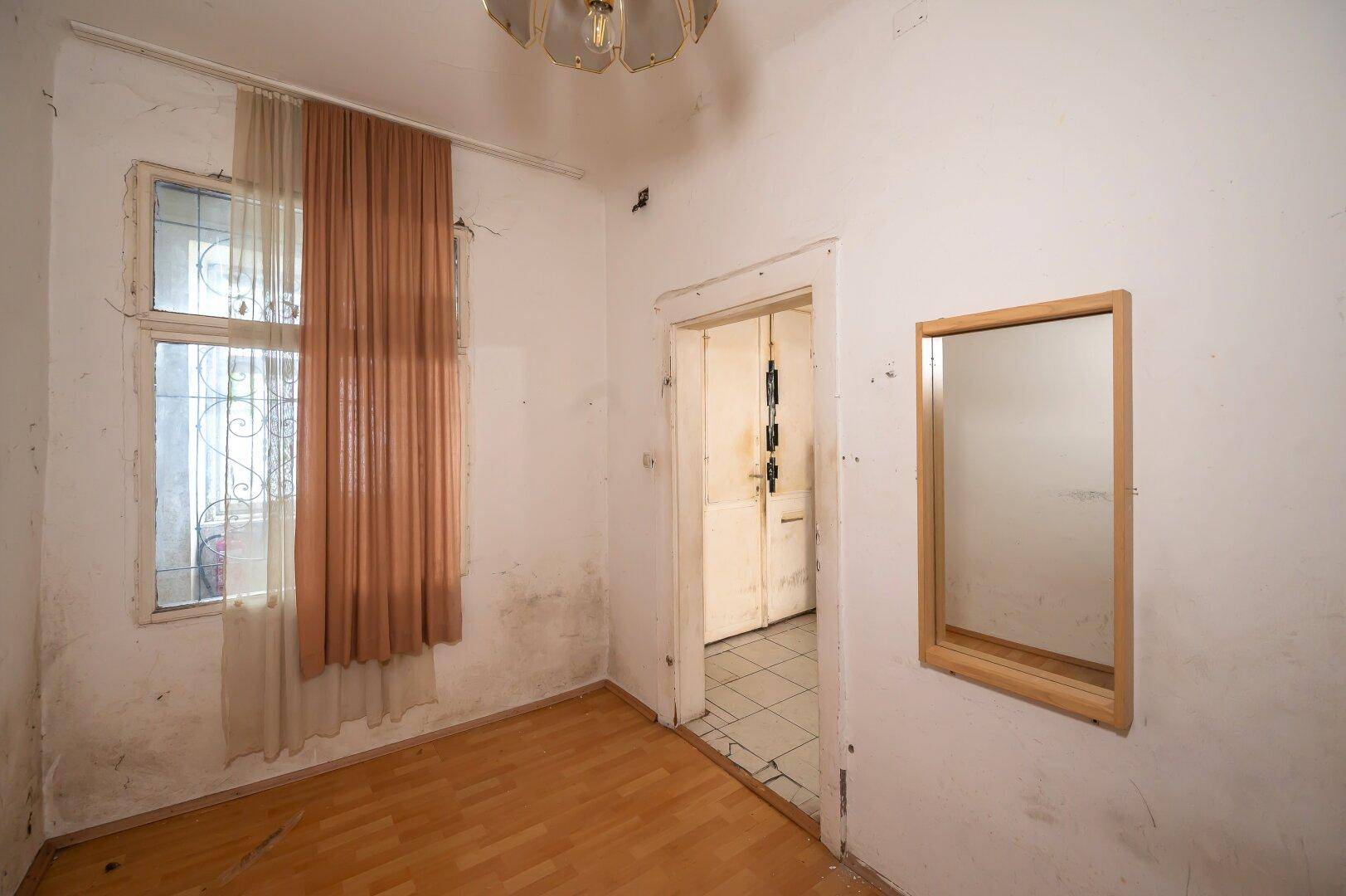 Helles Zimmer mit Holzboden, Fenster und einem Wandspiegel, das einen einfachen Eindruck vermittelt.