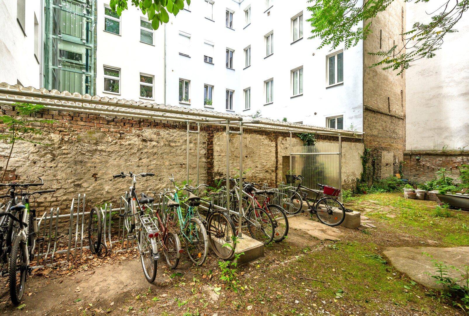 Innenhof mit Fahrradständern, Grünflächen und einem modernen Gebäude mit Glasaufzug im Hintergrund.