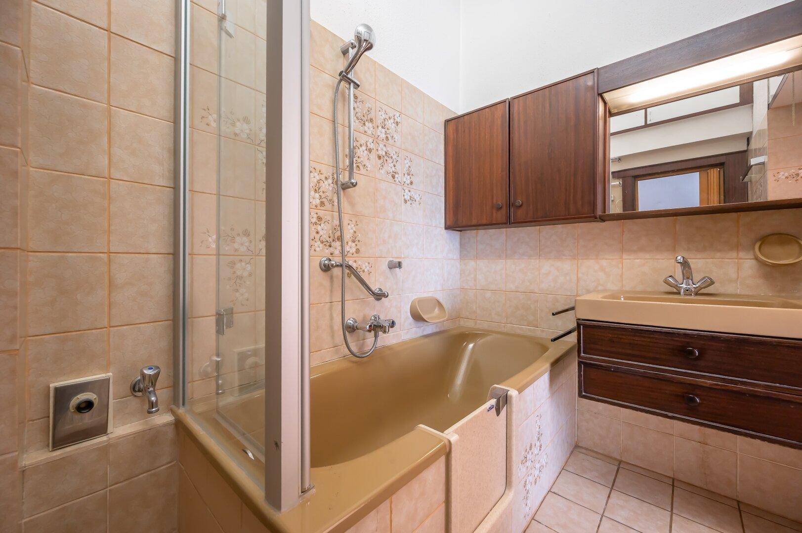 Badezimmer mit Badewanne und Duschabtrennung, ausgestattet mit beigen Fliesen im 80er-Jahre-Stil.