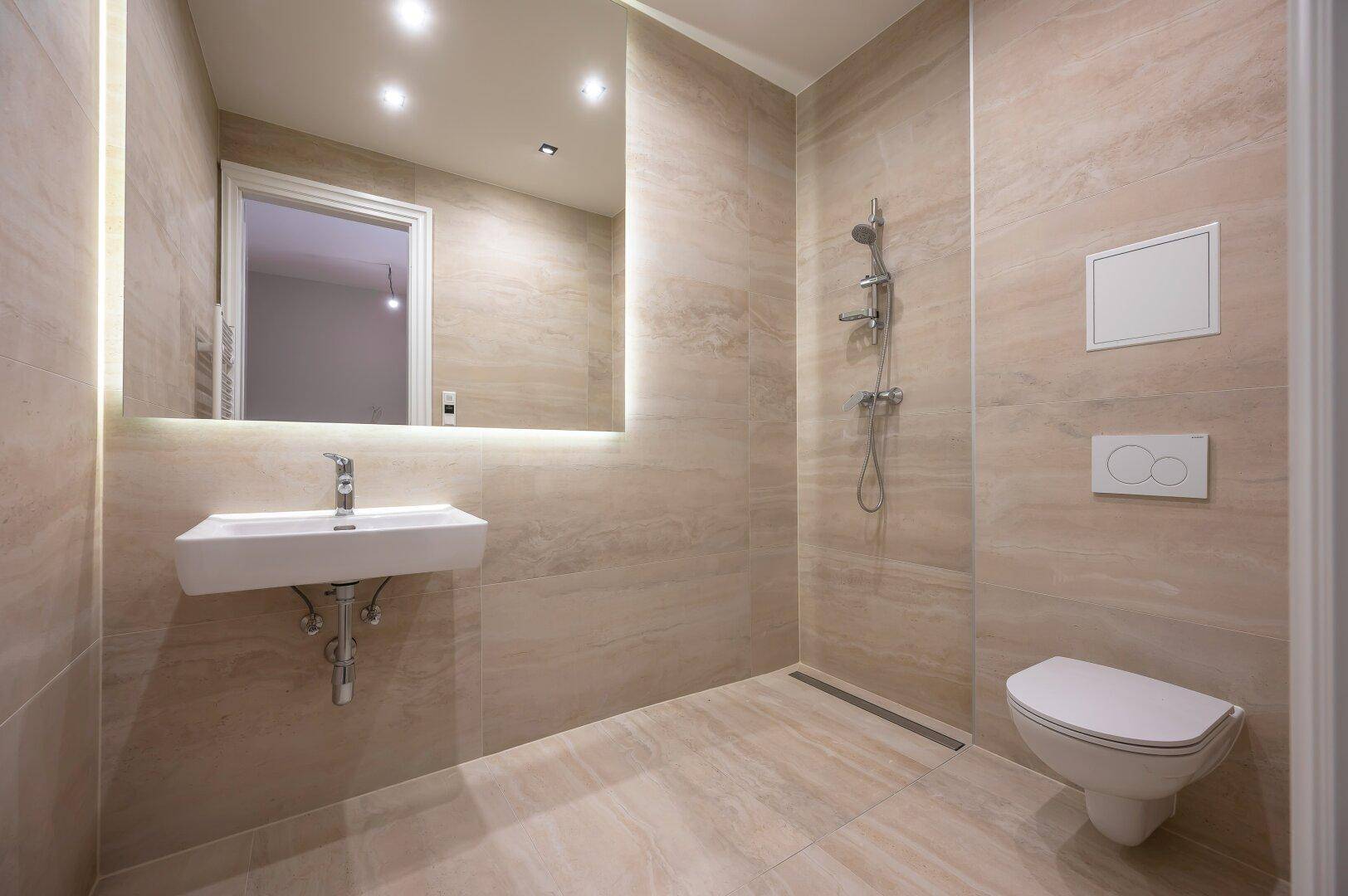 Modernes Badezimmer mit beigen Fliesen, wandhängendem WC und begehbarer Dusche, beleuchtet durch einen Spiegelschrank.