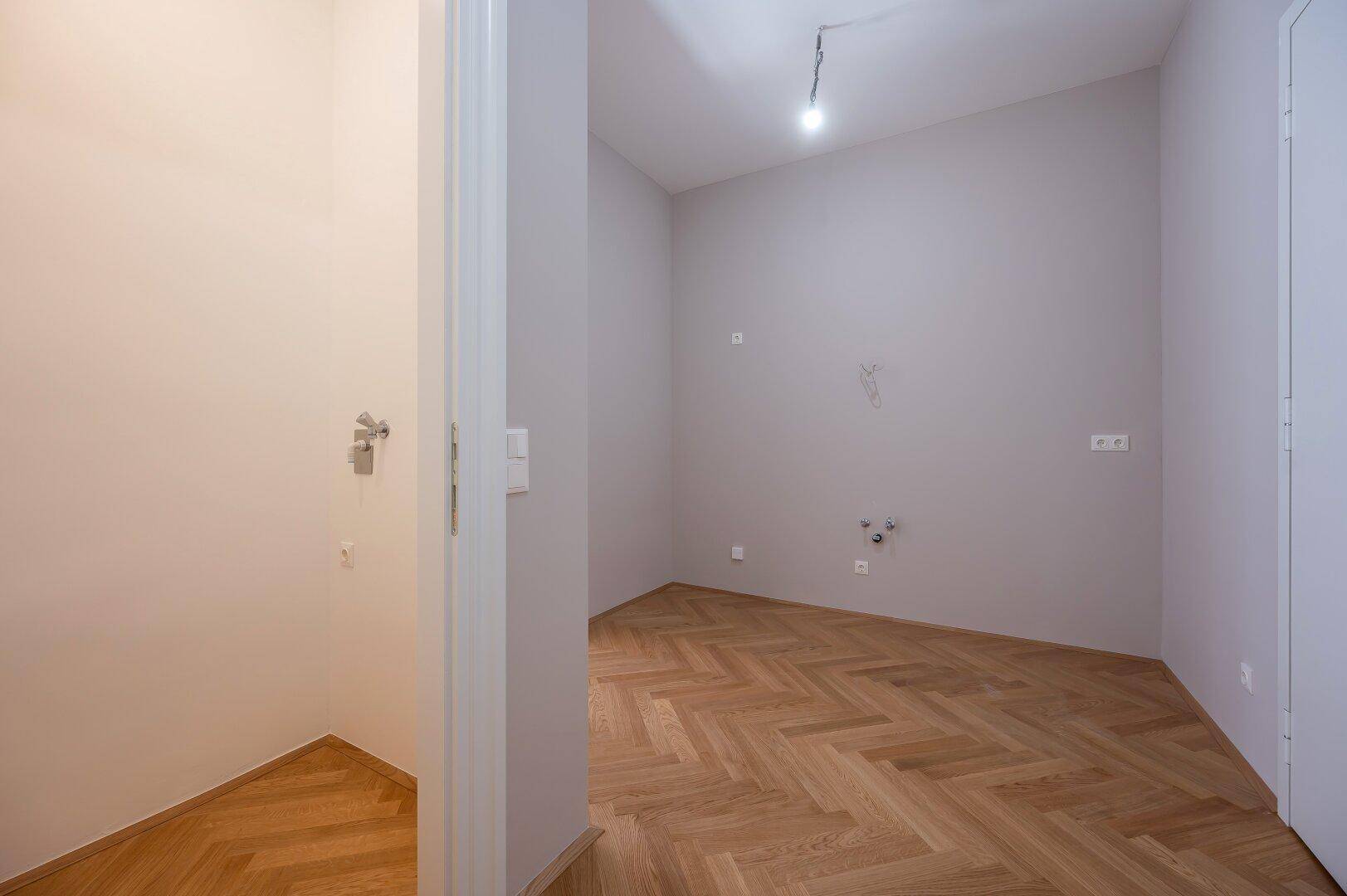 Leerer Raum mit Fischgrätparkettboden und Anschlüssen für Sanitärinstallationen, ideal als Hauswirtschaftsraum.