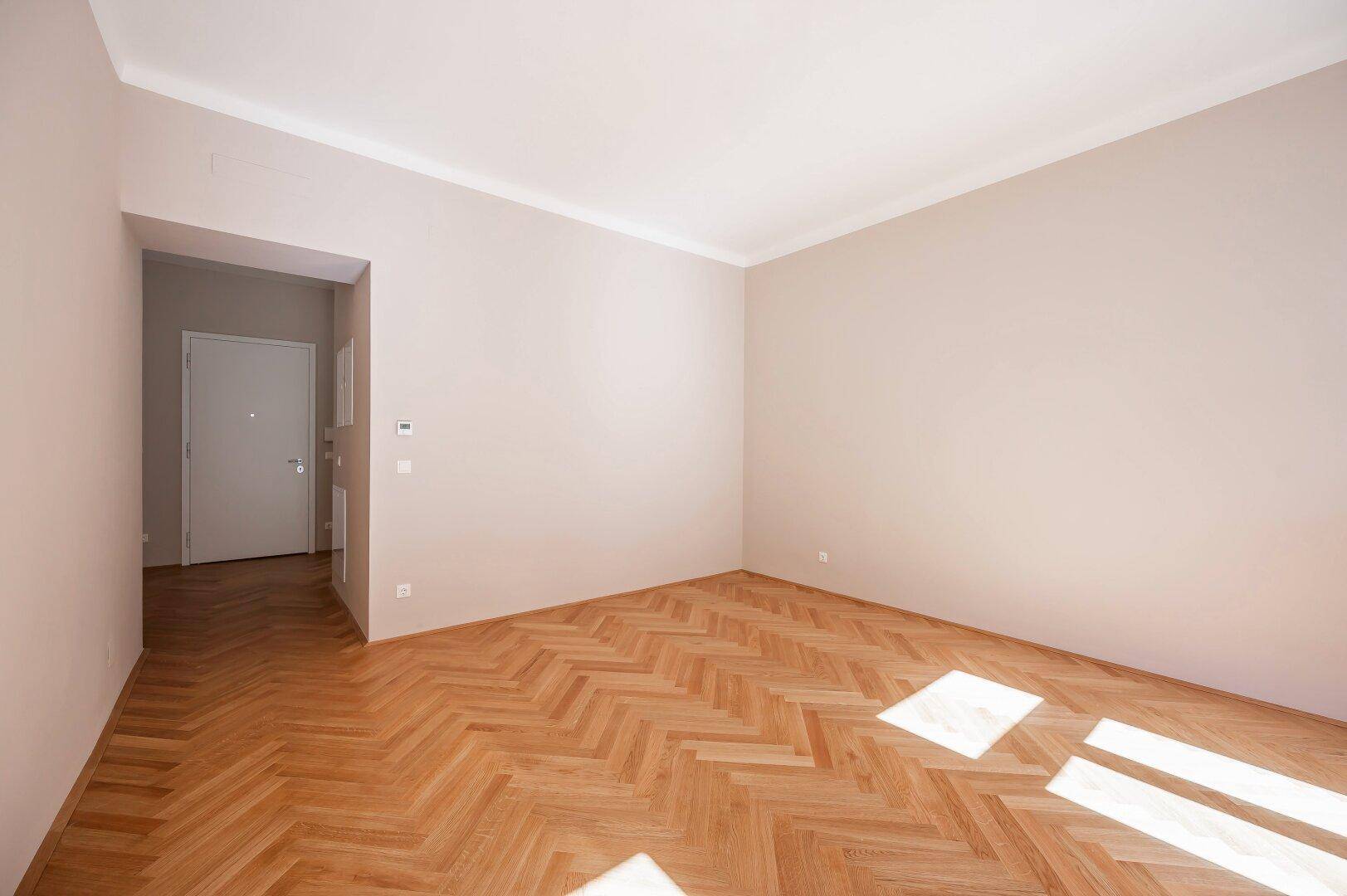 Geräumiges Zimmer mit Fischgrätparkettboden und Zugang zu einem Flur mit Eingangstür.