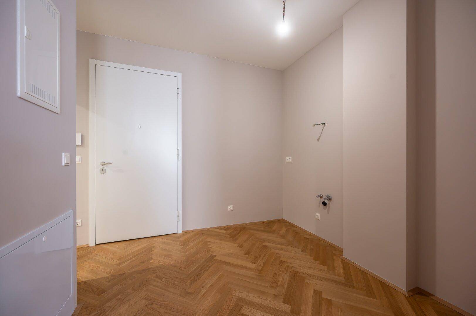 Eingangsbereich mit Fischgrätparkett, weißer Eingangstür und Anschlüssen für eine Waschmaschine.