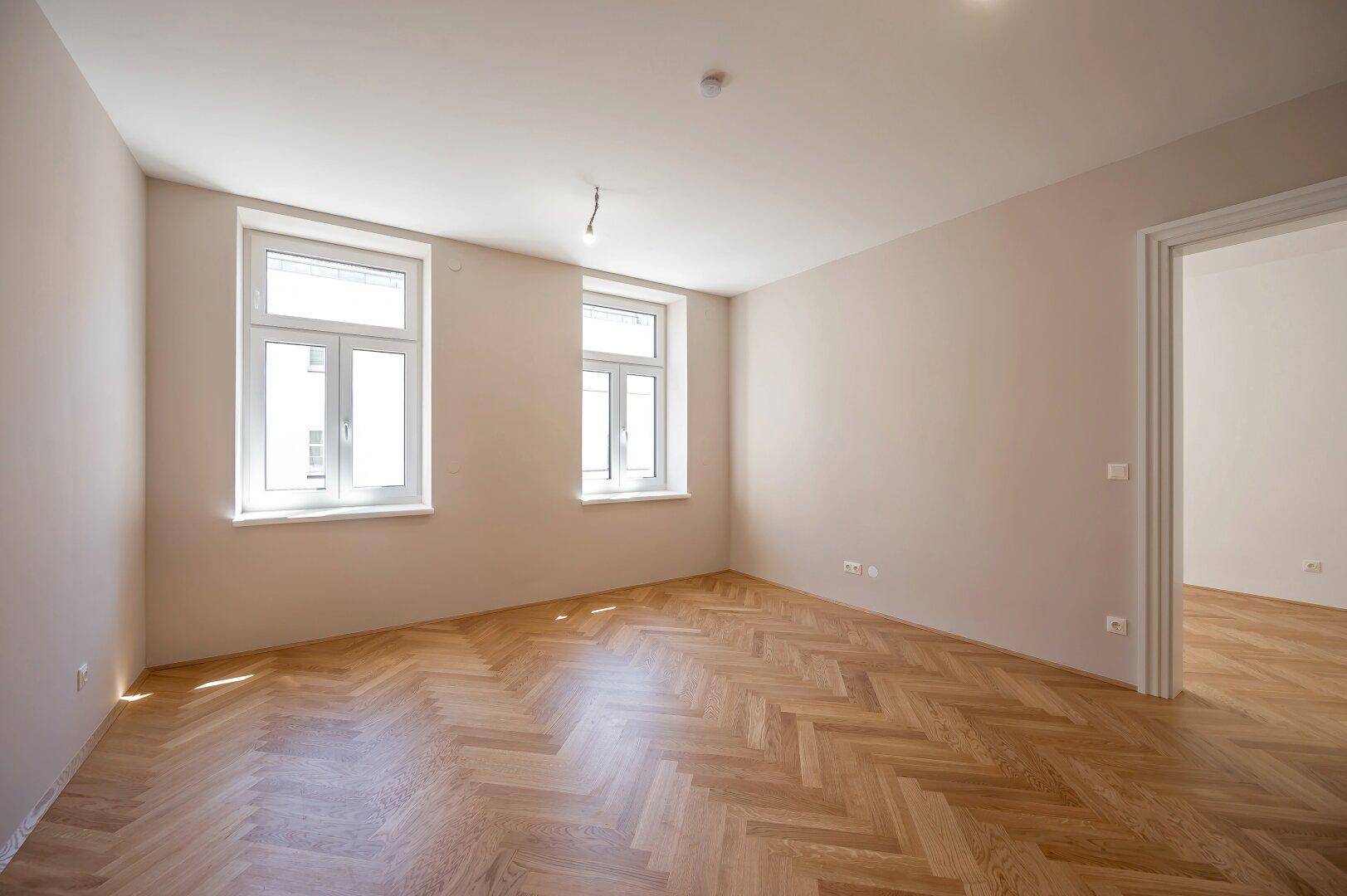Heller, leerer Raum mit zwei Fenstern und Fischgrätparkett, ideal für Wohn- oder Schlafbereich.