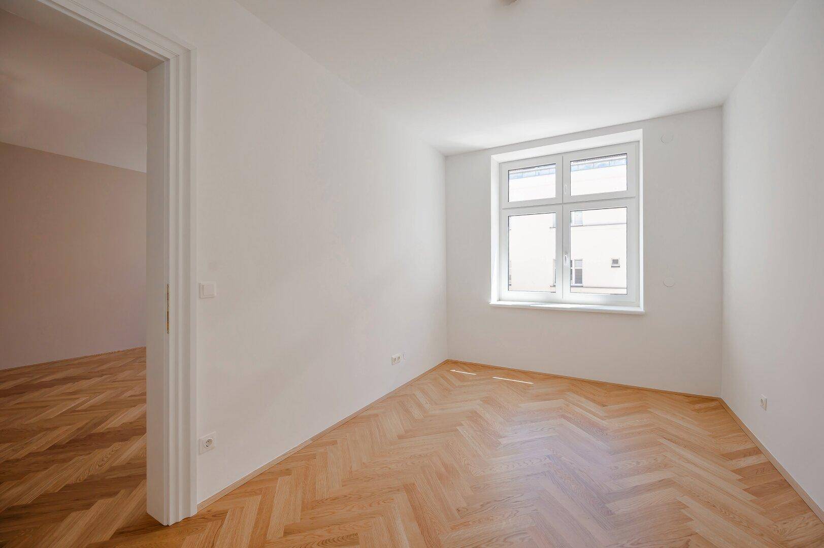 Leerer Raum mit Fischgrätparkettboden und einem großen Fenster, bereit zur individuellen Gestaltung.