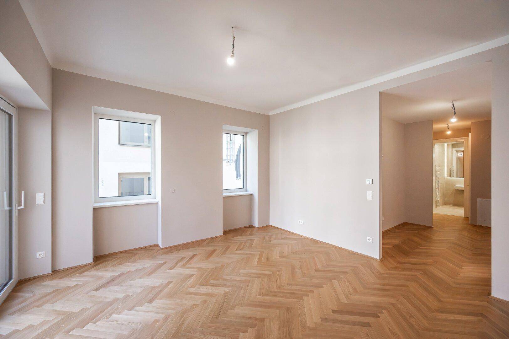 Geräumiges Zimmer mit elegantem Fischgrätparkett und Blick in den Flur mit angrenzendem Badezimmer.
