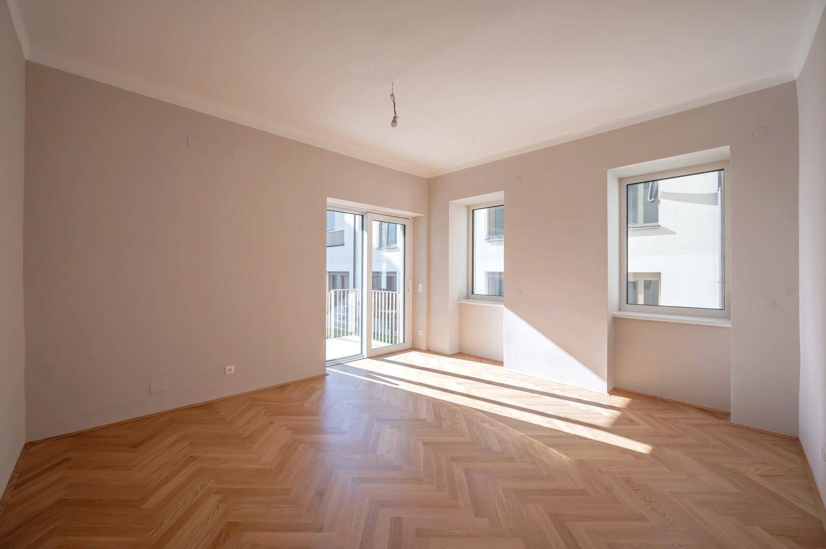 Helles Zimmer mit Fischgrätparkett und Balkonzugang, durchflutet von Sonnenlicht, ideal für Wohnzwecke.