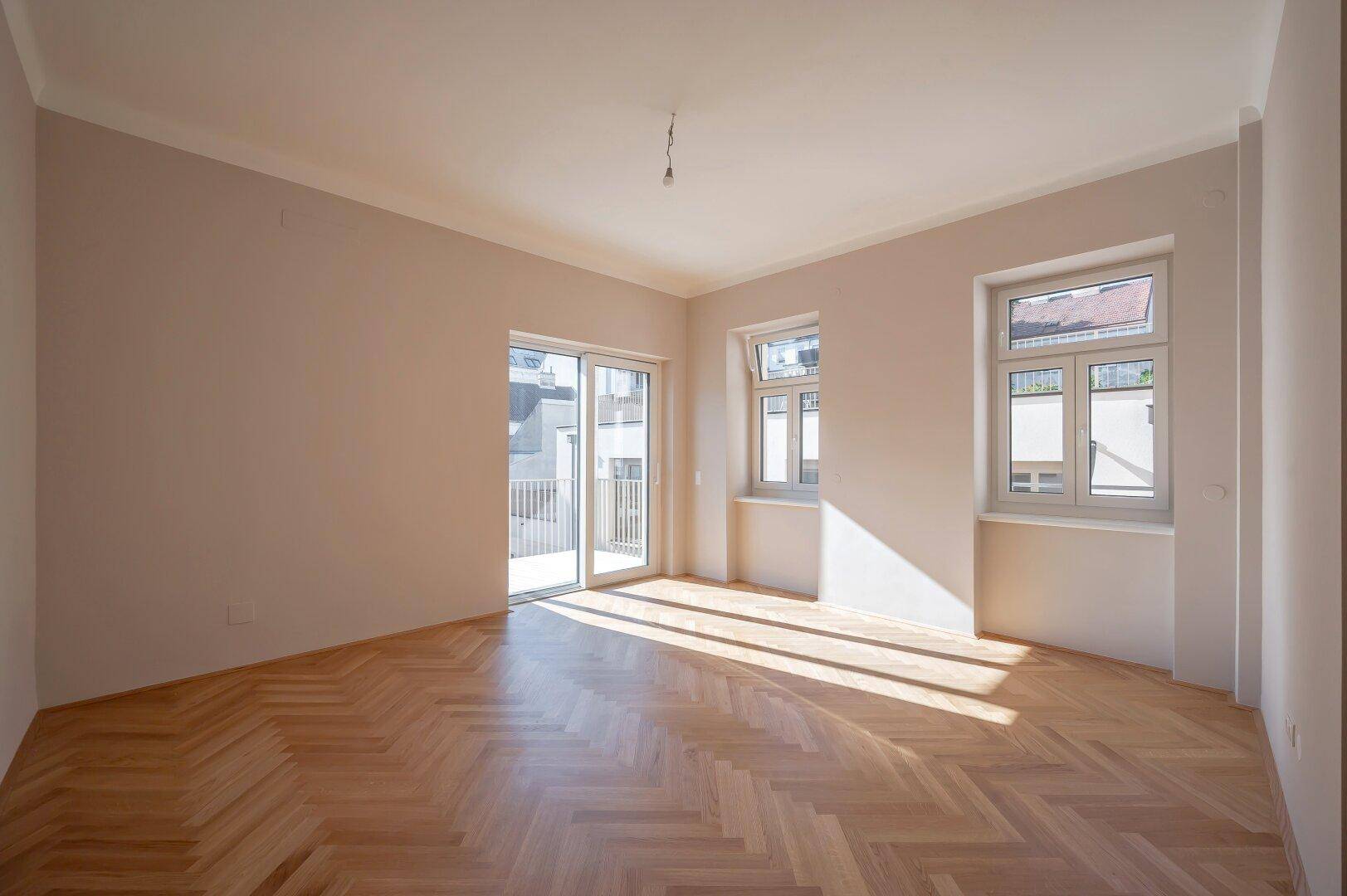 Heller Wohnraum mit Fischgrätparkett, großen Fenstern und Zugang zu einem Balkon.