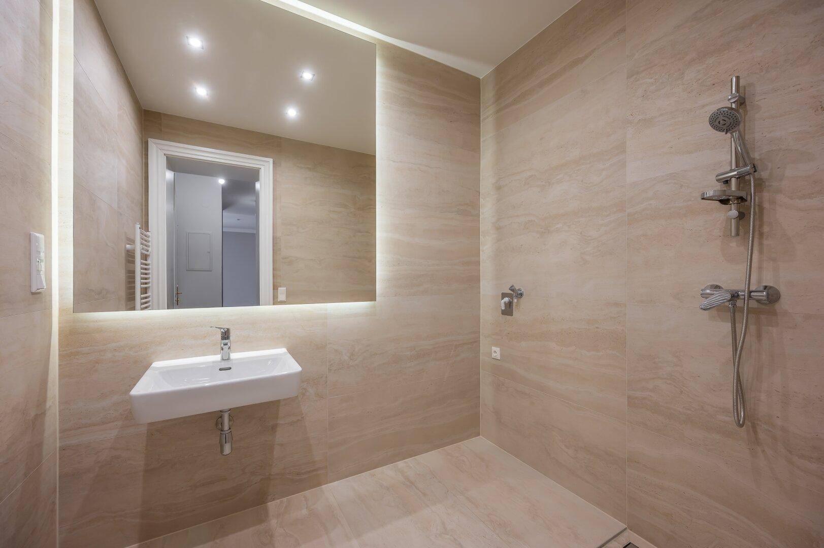 Modernes Badezimmer mit beigefarbenen Fliesen, bodengleicher Dusche und beleuchtetem Spiegel.
