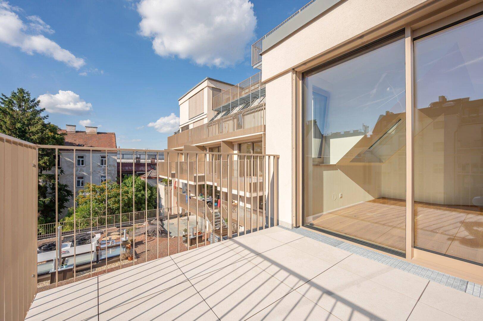 Sonniger Balkon mit Fliesenboden und Blick auf die umliegenden Gebäude und den Himmel.