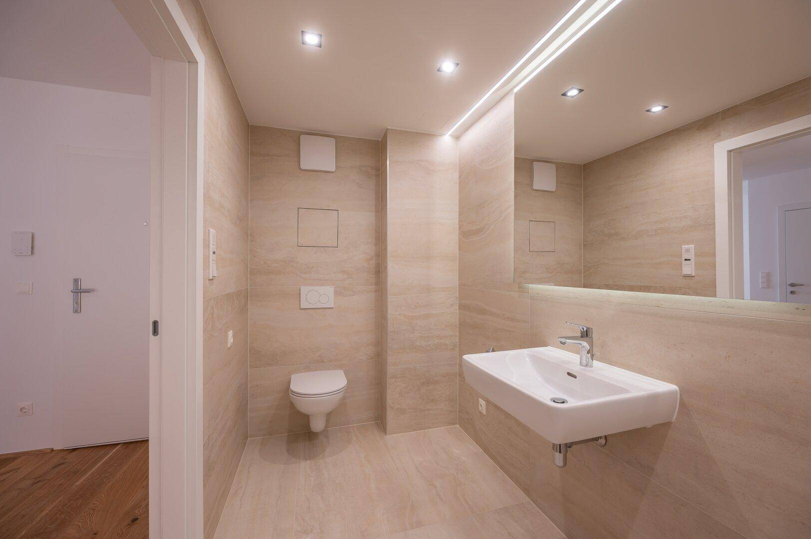 Modernes Badezimmer mit beigefarbenen Fliesen, Wand-WC und beleuchtetem Spiegel über dem Waschbecken.