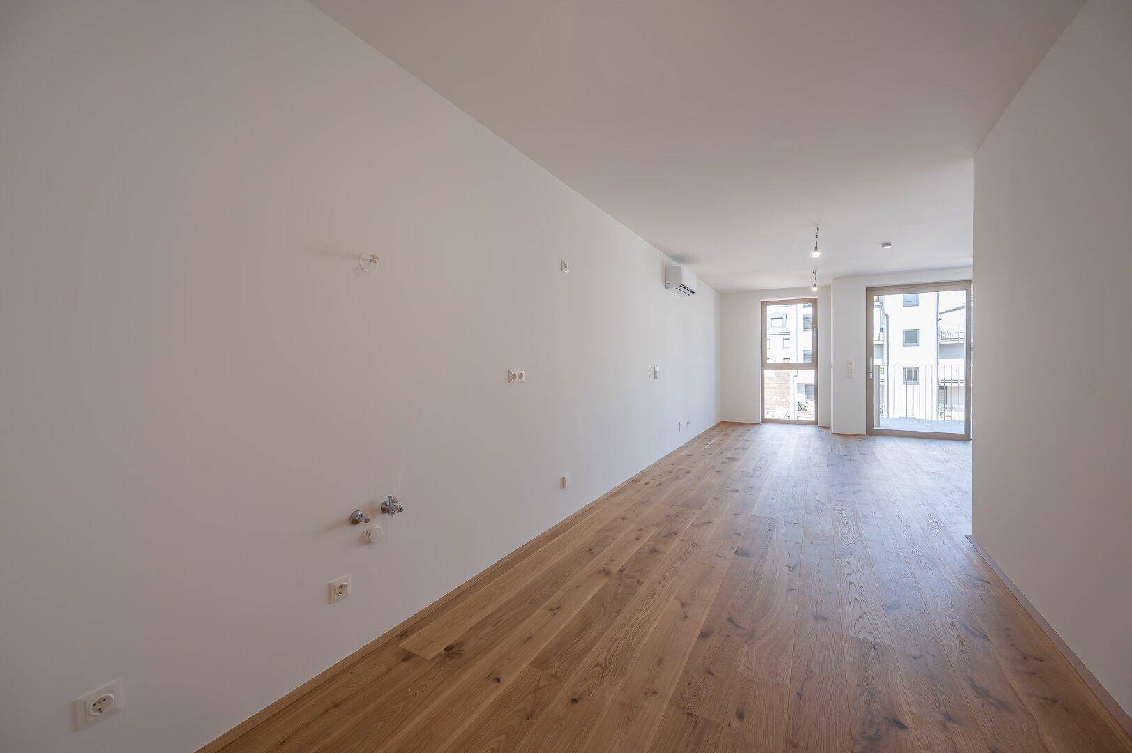 Heller Raum mit Holzboden, Klimaanlage und Zugang zu einem sonnigen Balkon.