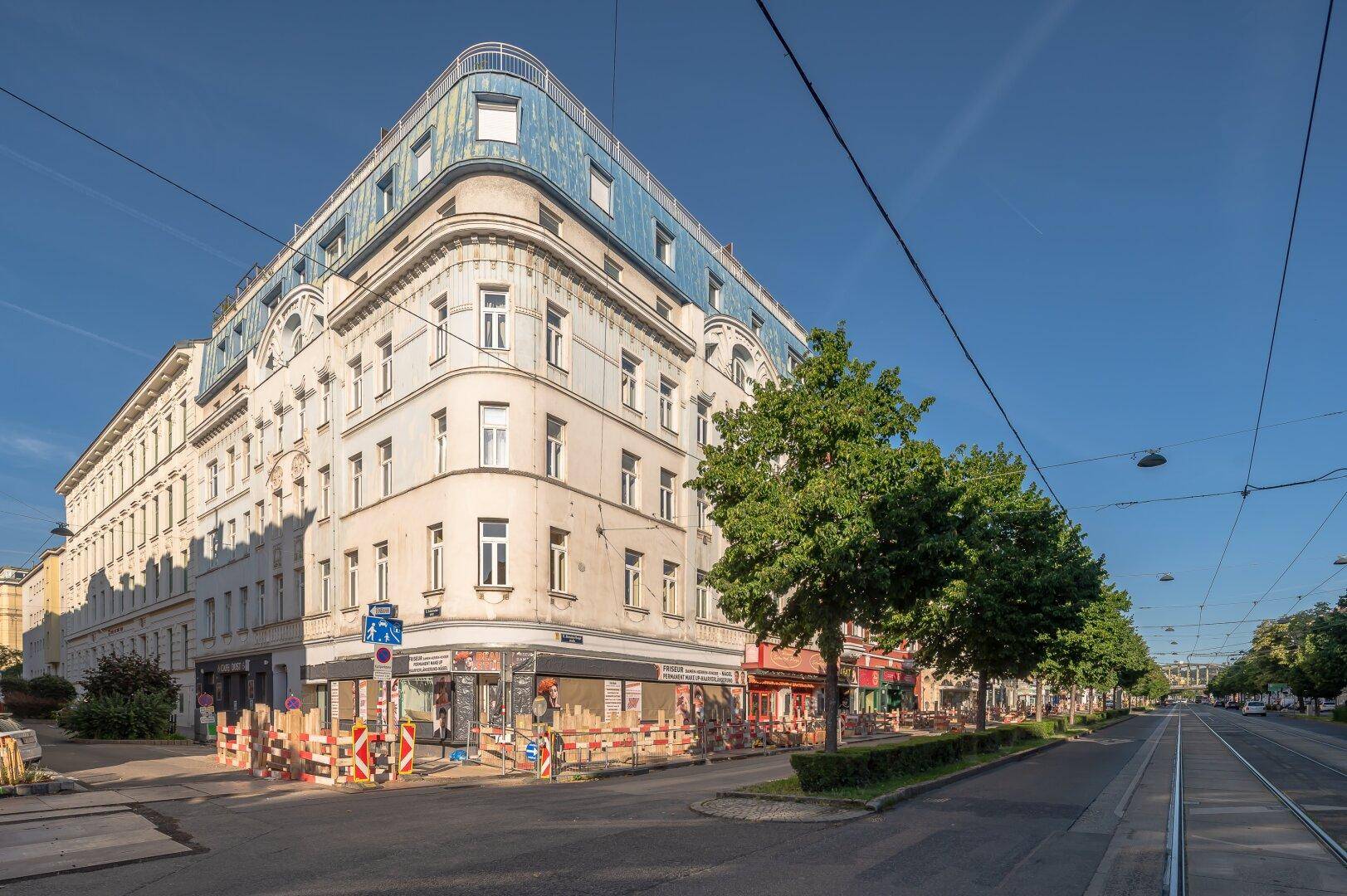 Eckgebäude in einer städtischen Umgebung mit Geschäften im Erdgeschoss und blauer Dachkonstruktion.