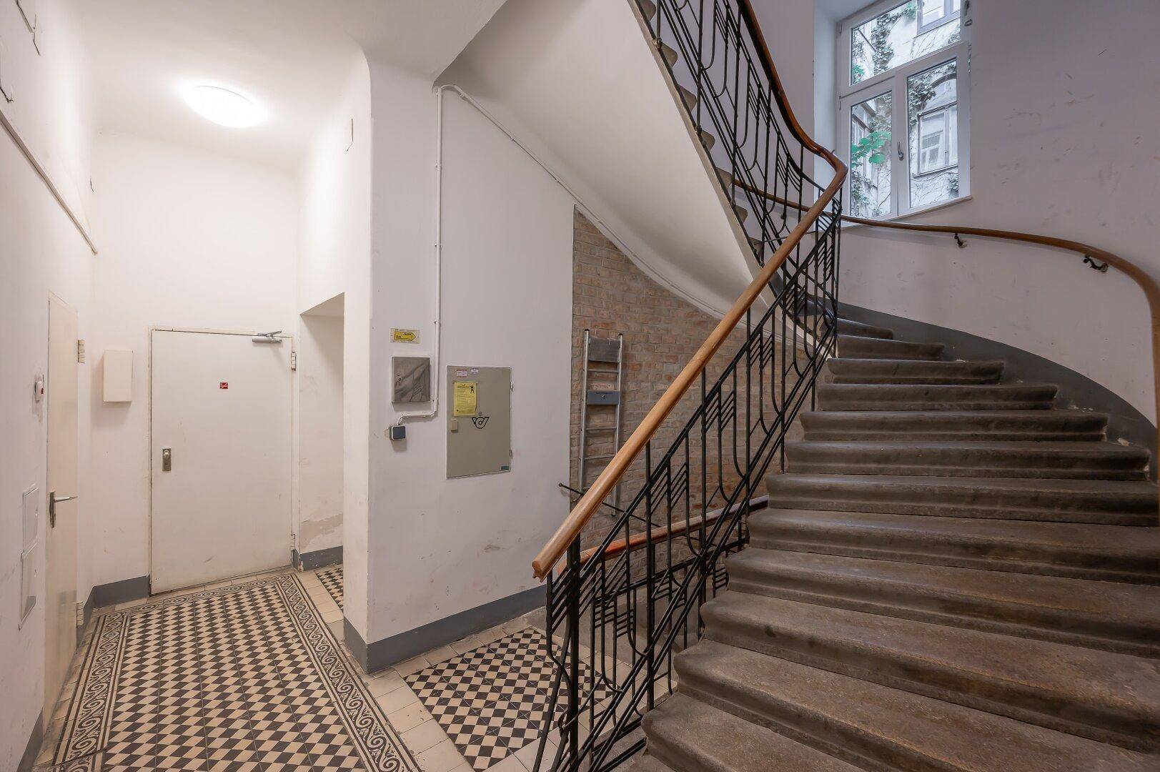 Treppenhaus in einem Altbau mit geschwungenem Geländer und gemustertem Fliesenboden im Eingangsbereich.