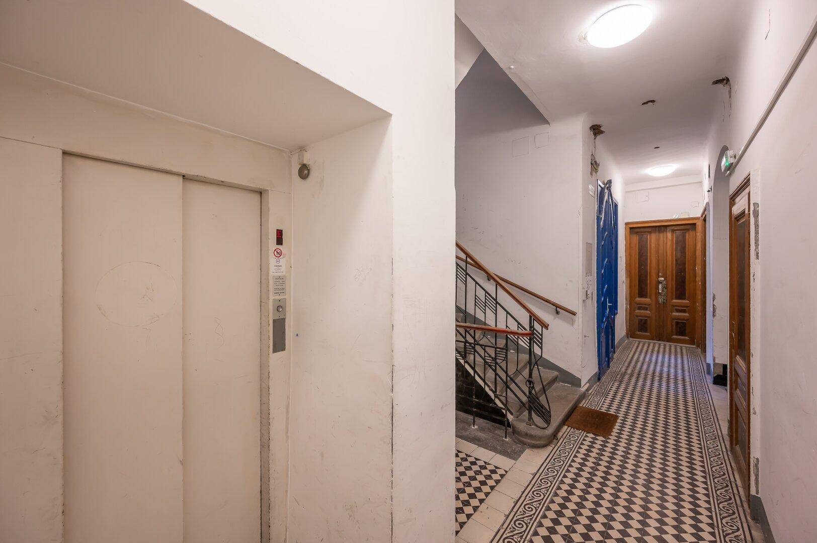 Gemeinschaftsflur in einem Altbau mit Aufzug, Treppenhaus und gemustertem Fliesenboden.