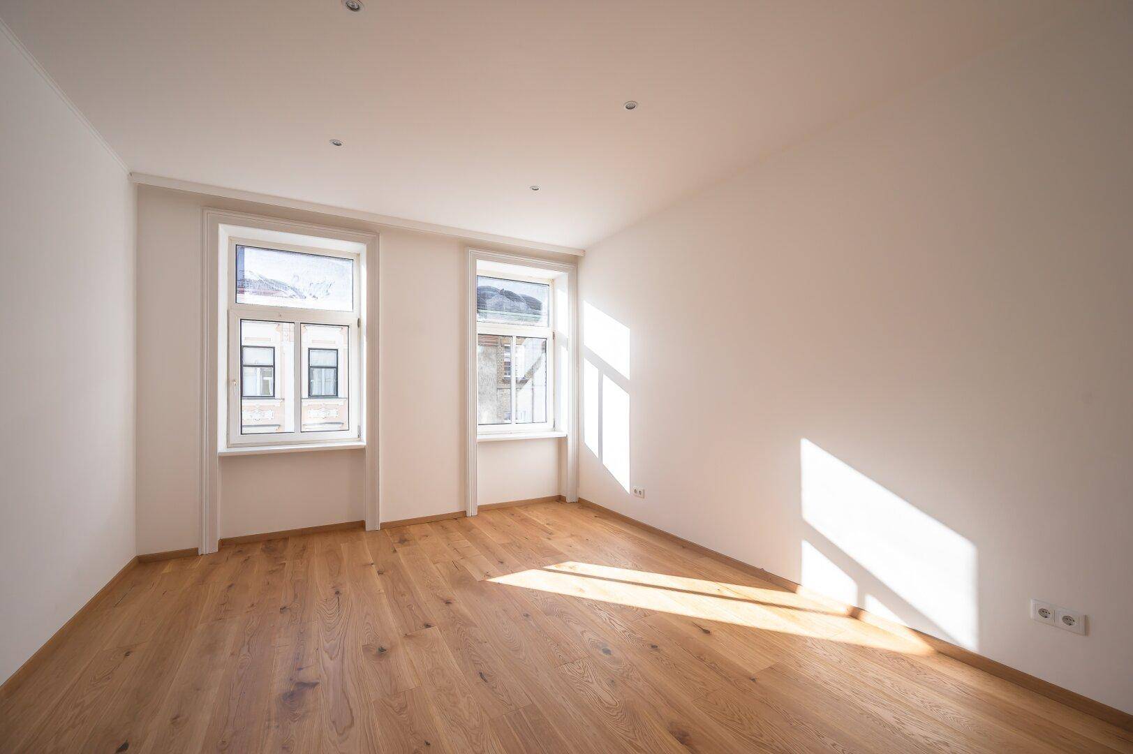 Heller, leerer Raum mit Holzboden und zwei Fenstern, die viel Sonnenlicht hereinlassen.