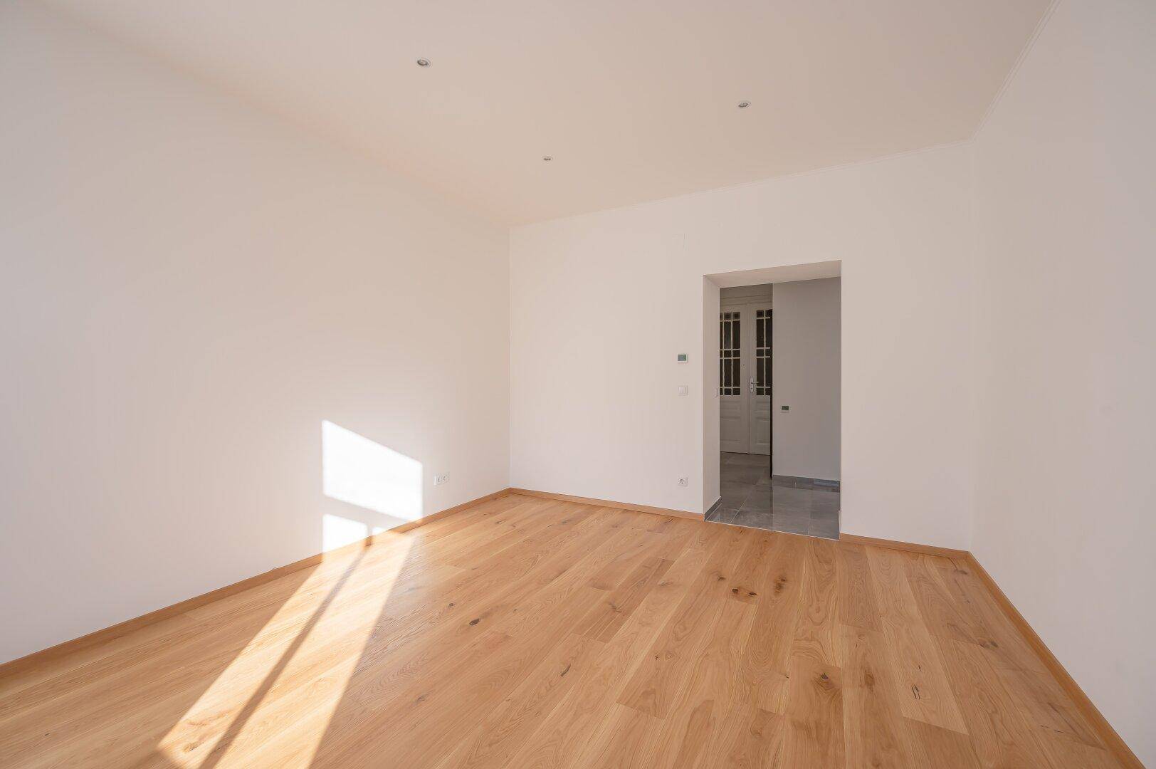 Geräumiger, leerer Raum mit Holzboden und offenem Durchgang zu einem gefliesten Bereich.