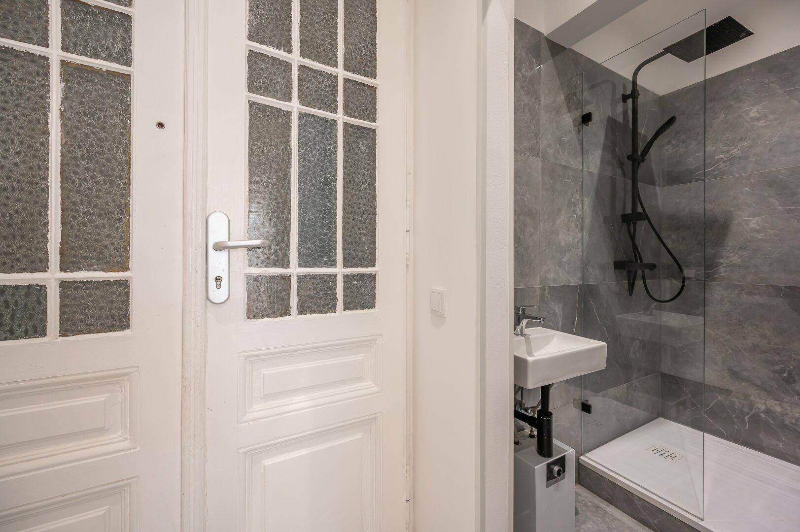 Detailansicht einer modernen Dusche mit grauen Fliesen und kleinem Waschbecken neben einer traditionellen Tür.