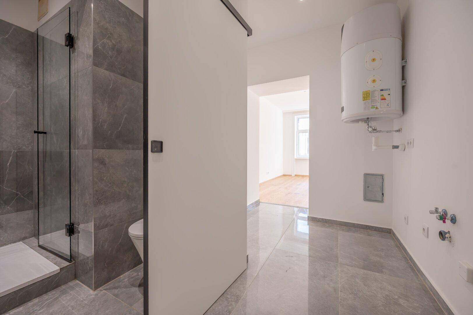 Flur mit grauen Fliesen, Warmwasserbereiter und Blick auf eine moderne Dusche.