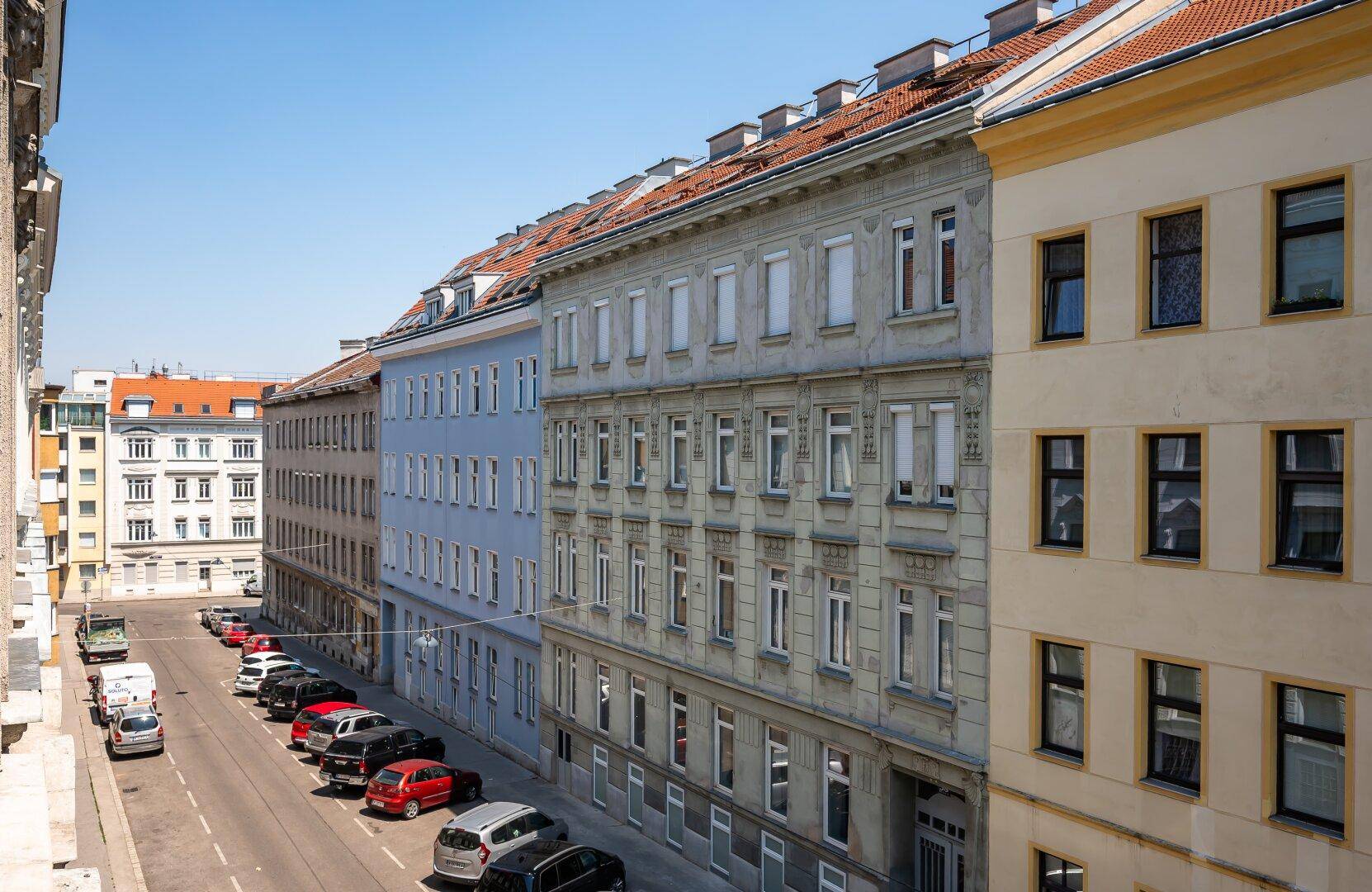 Lange Reihe von Altbauten entlang einer sonnigen Stadtstraße mit parkenden Autos.